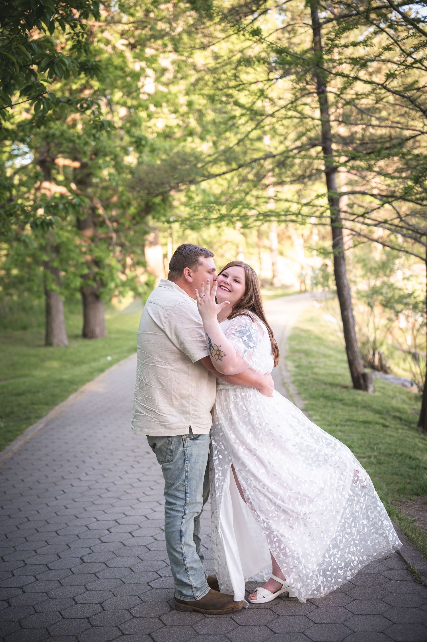 Couple sharing a romantic moment on a tree-lined brick pathway, woman in white lace dress laughing joyfully.