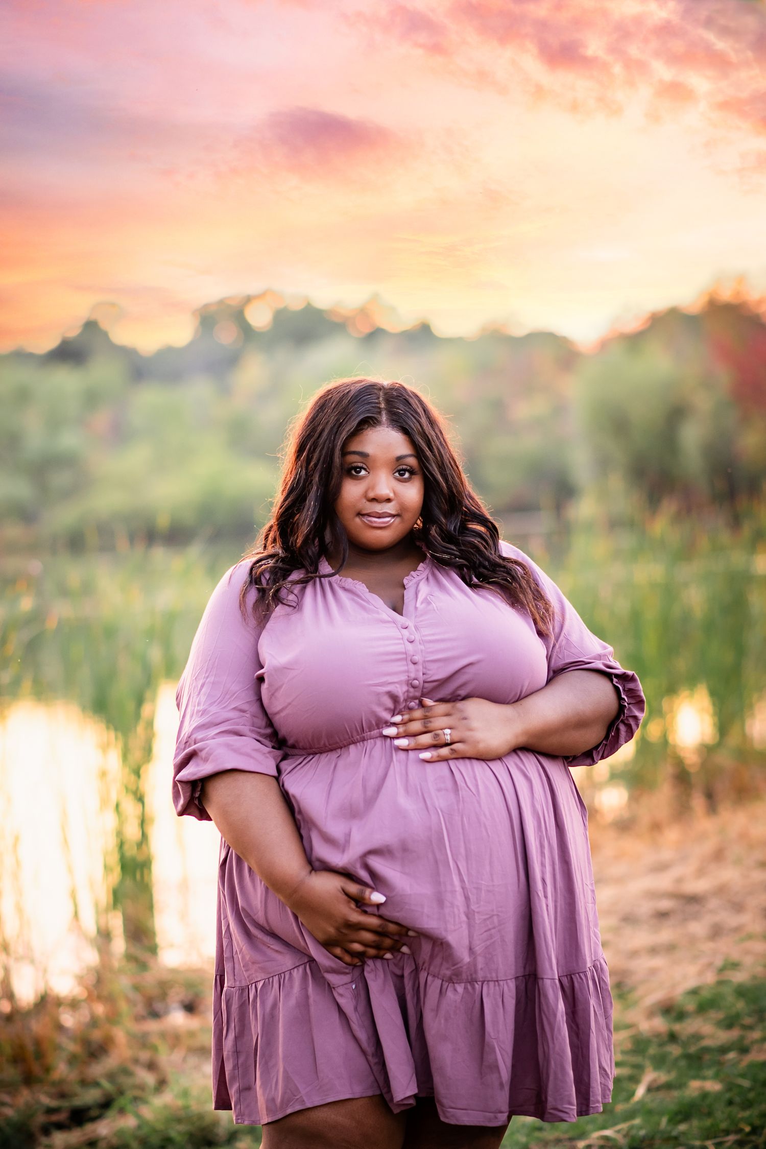 A person in a purple dress poses for a maternity photo at sunset by a lake.