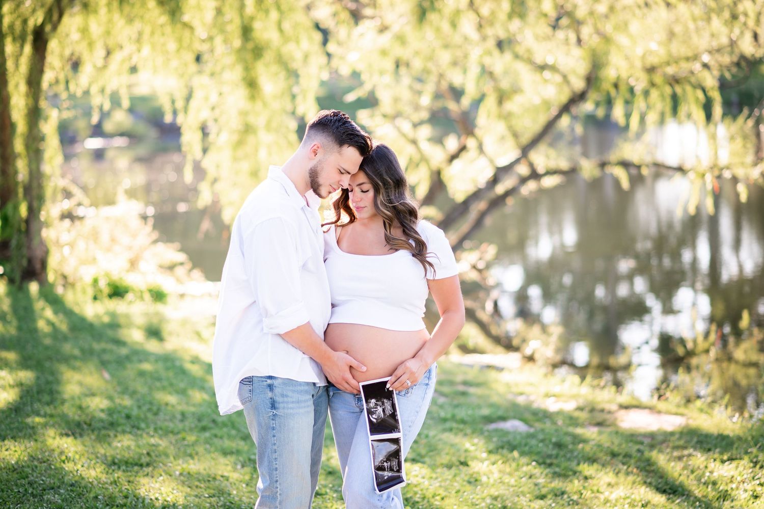 Couple holding ultrasound photos during outdoor maternity photoshoot by a pond, surrounded by lush green trees.