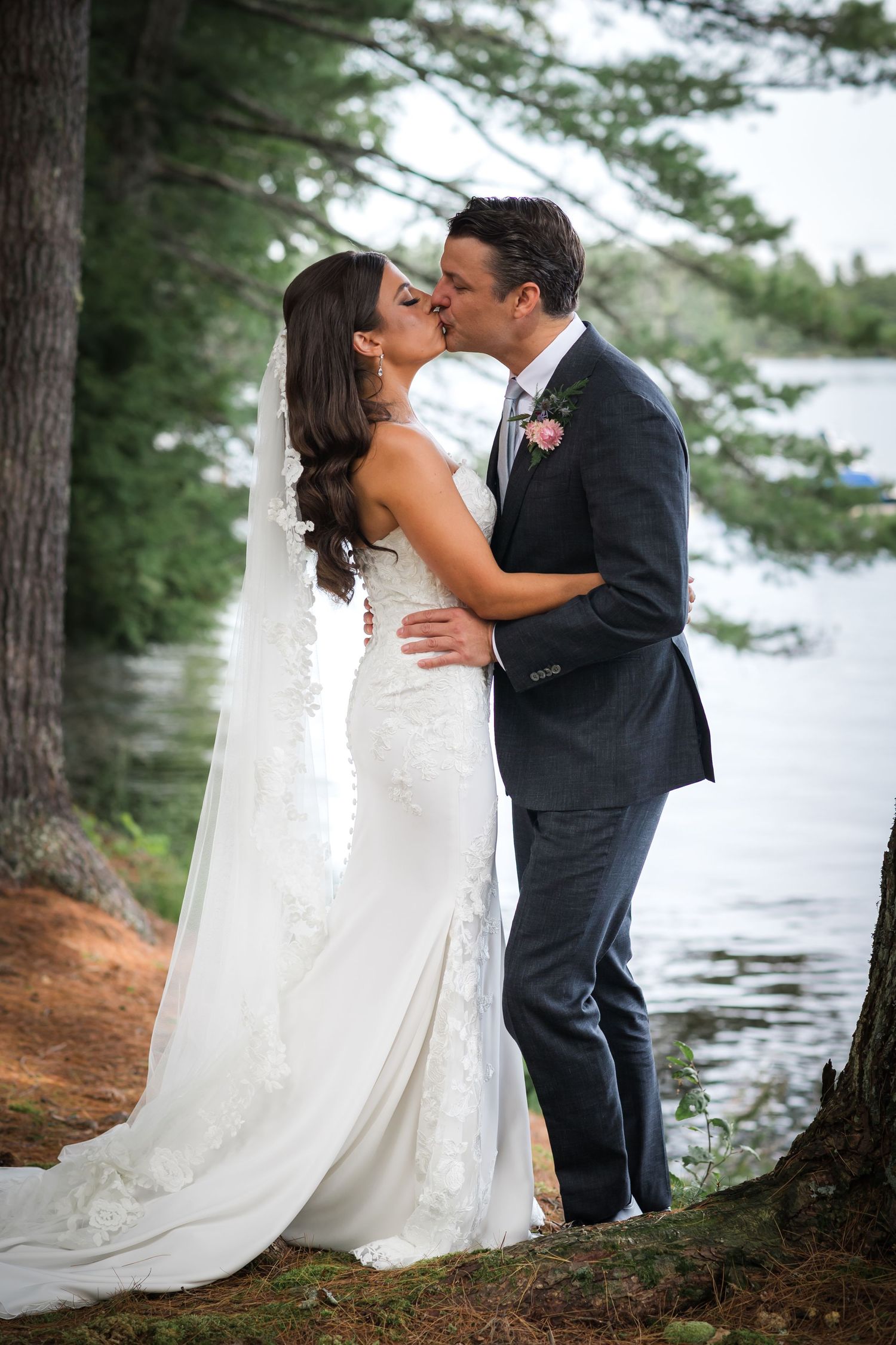 A romantic wedding couple shares a kiss by a lakeside surrounded by tall pine trees on a tranquil afternoon.