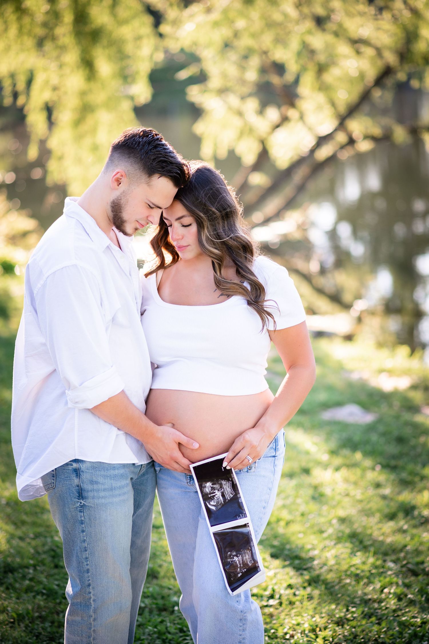 Expecting couple in white tops and jeans holding ultrasound photos outdoors, touching foreheads tenderly near a sunlit pond.
