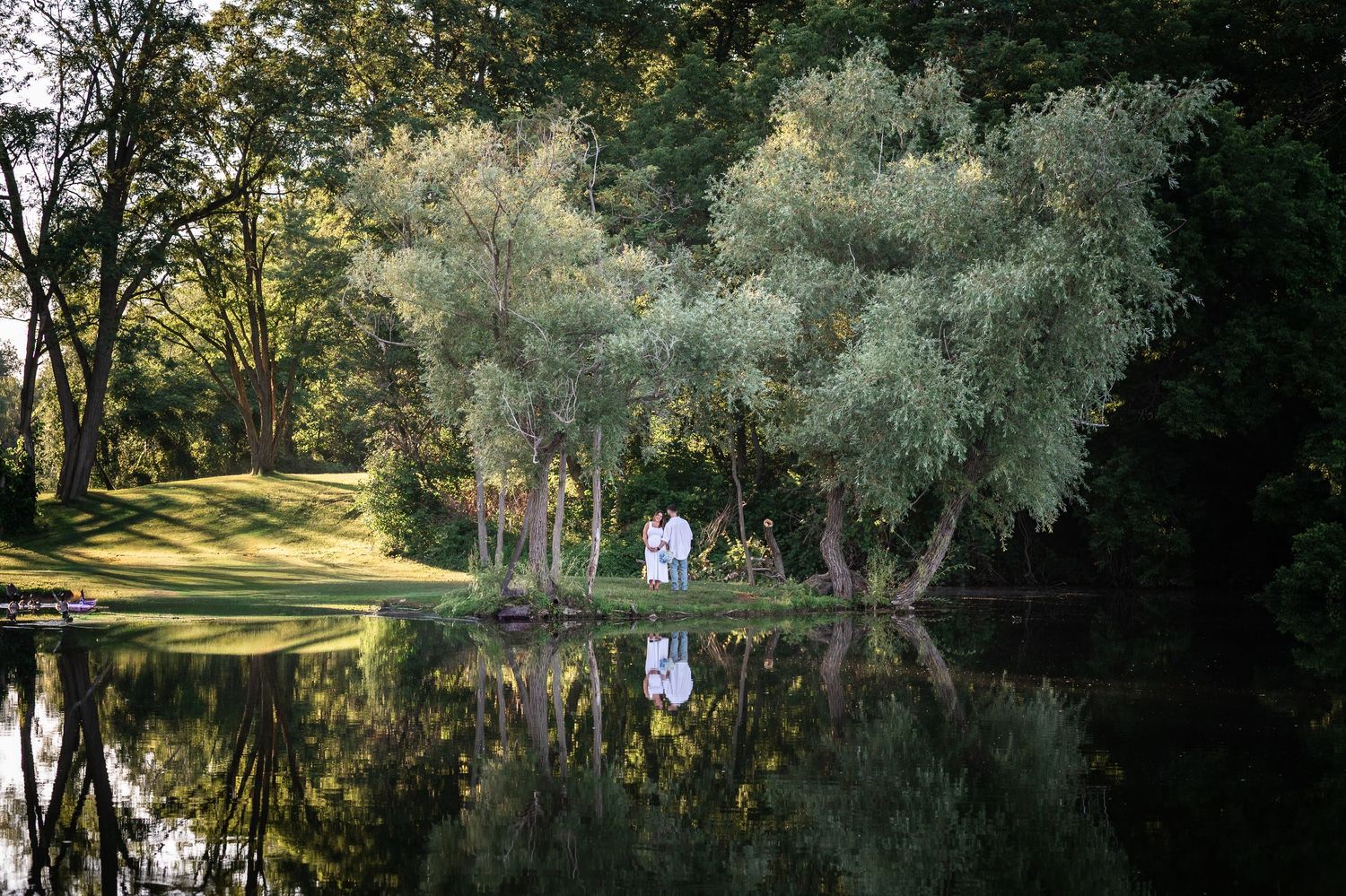 Couple standing under willow trees beside a tranquil reflective pond in a lush green park setting.