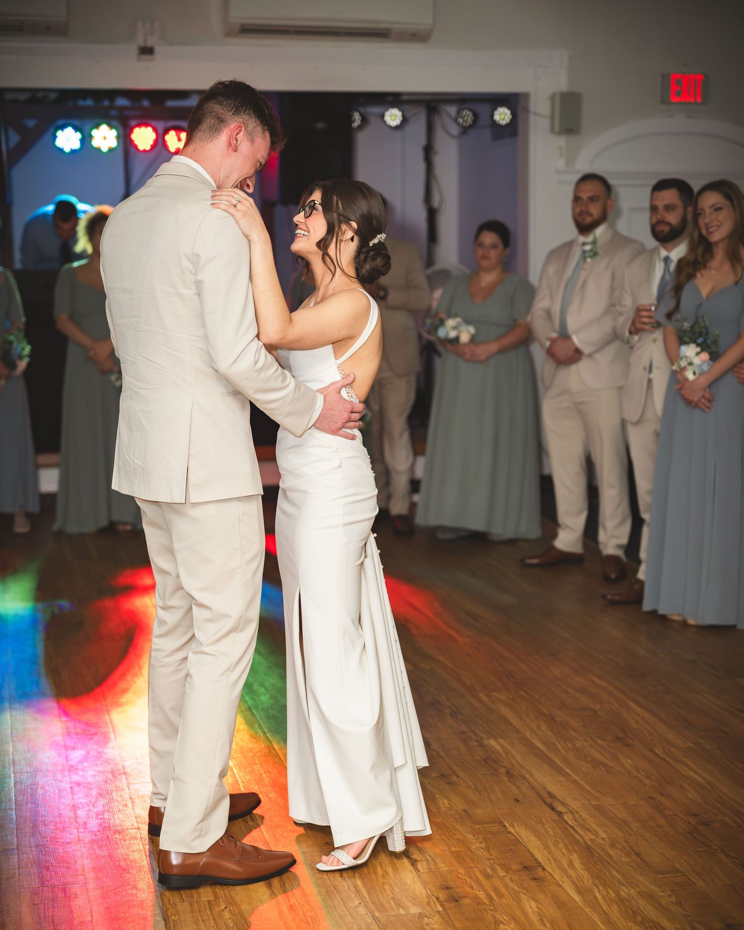 Bride and groom share their first dance at wedding reception, surrounded by guests in sage and tan attire.