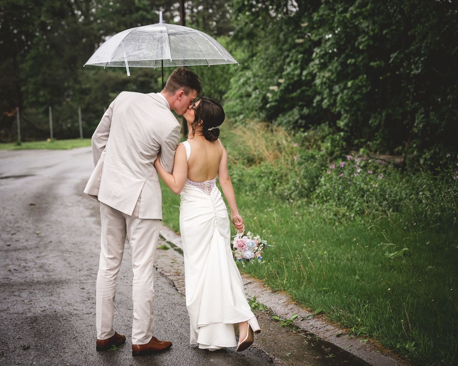 Bride and groom sharing a kiss under a clear umbrella on a rainy wedding day, bride holding a bouquet.
