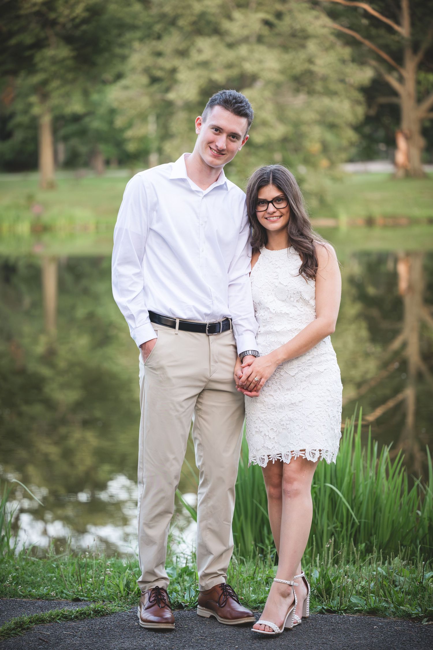 A smiling couple in white outfits holds hands near a serene pond surrounded by lush greenery.