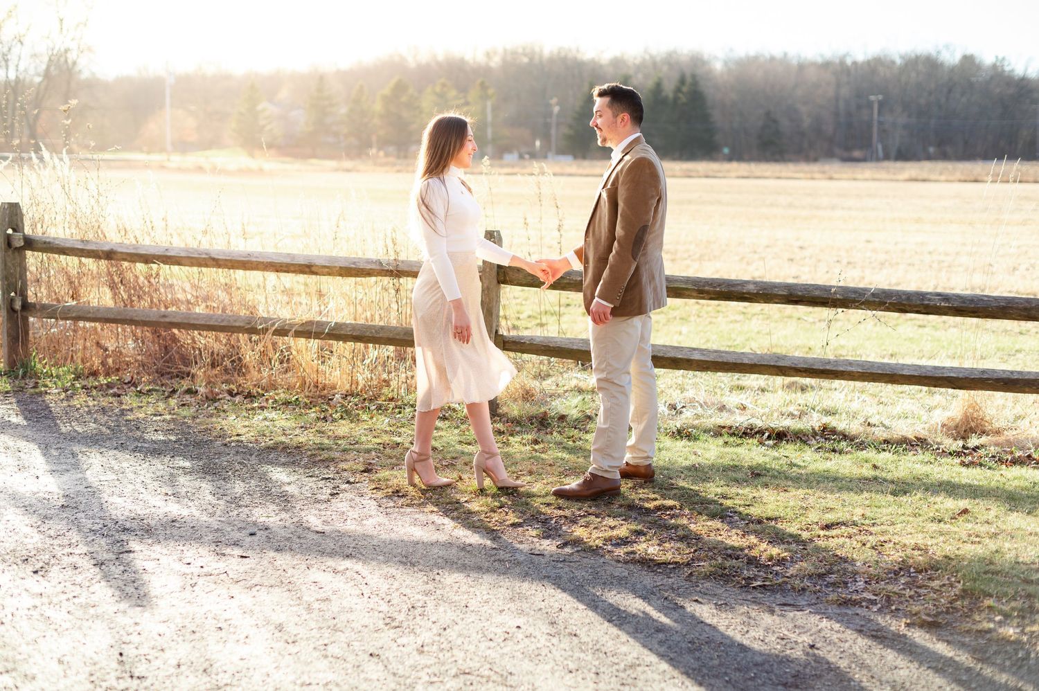 A romantic couple walks together along a rustic wooden fence at sunset in a pastoral countryside setting.