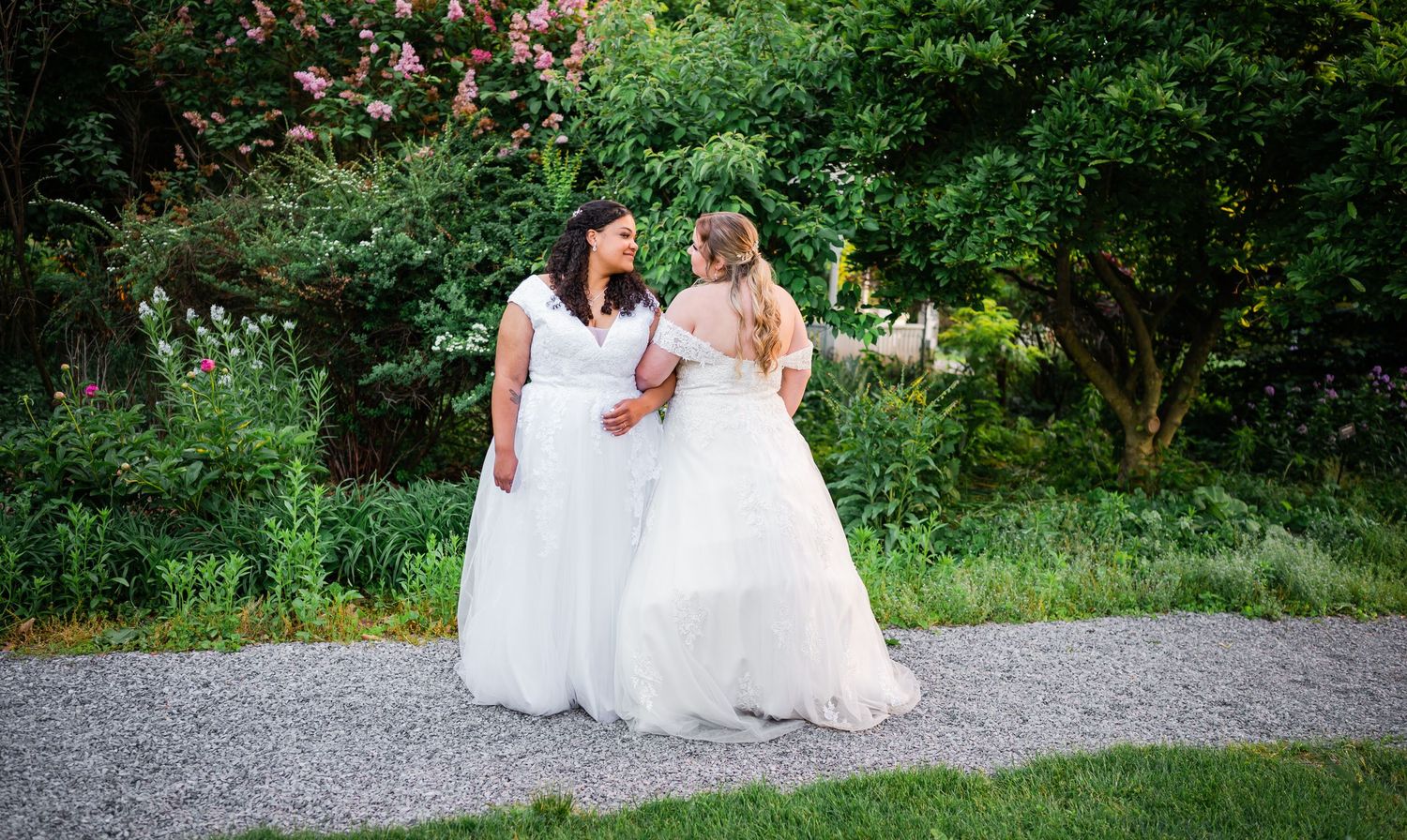 Two people in white wedding dresses share a joyful moment on a garden path.