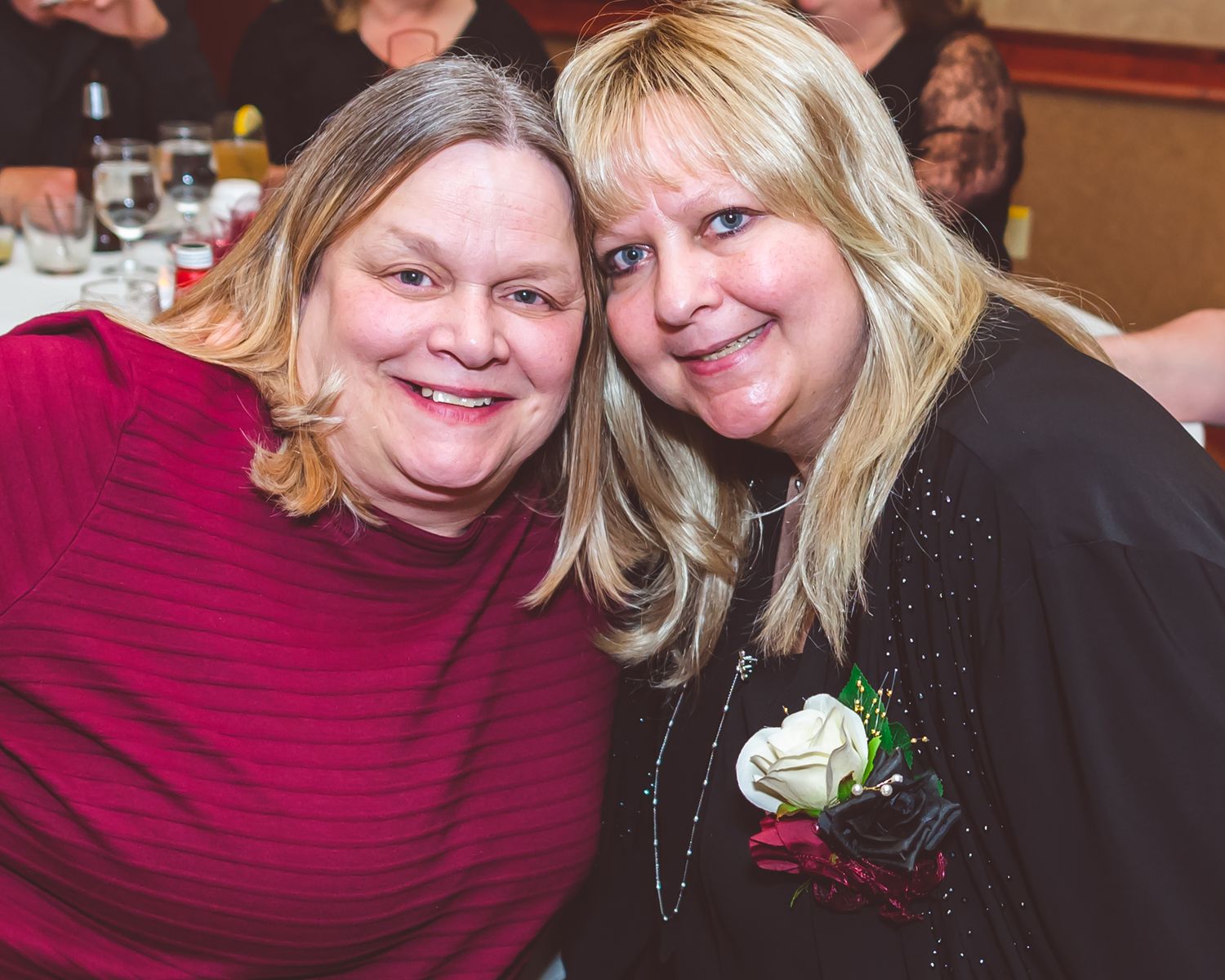 Two friends sharing a warm moment at a celebration event, one wearing red and one wearing black with a white corsage.