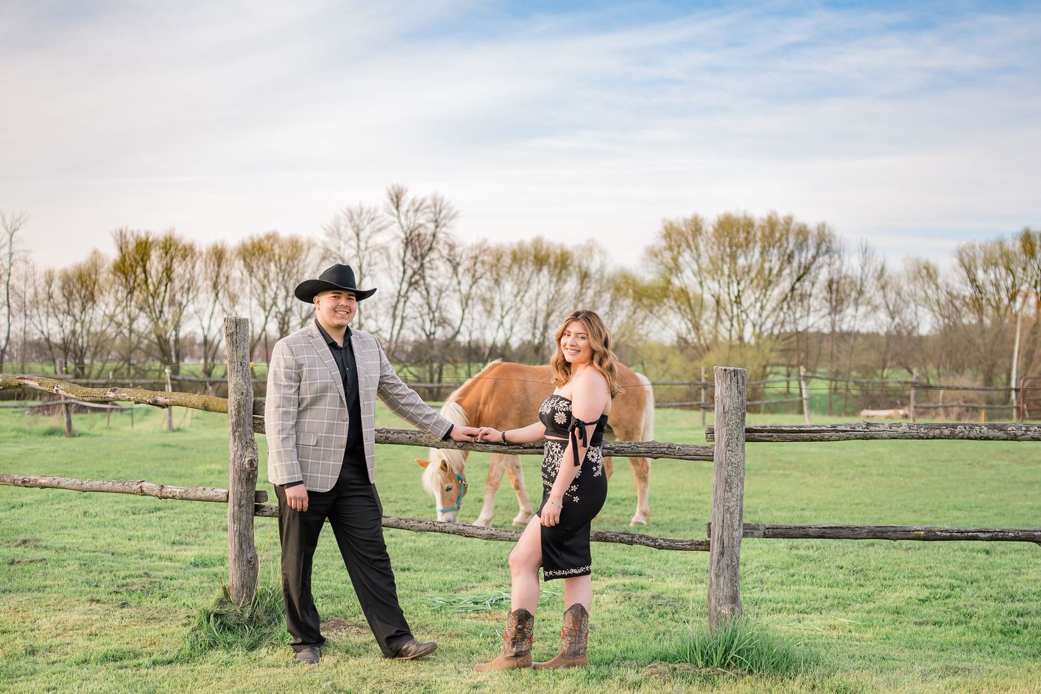 Country couple poses with horse in fenced pasture under spring sky.