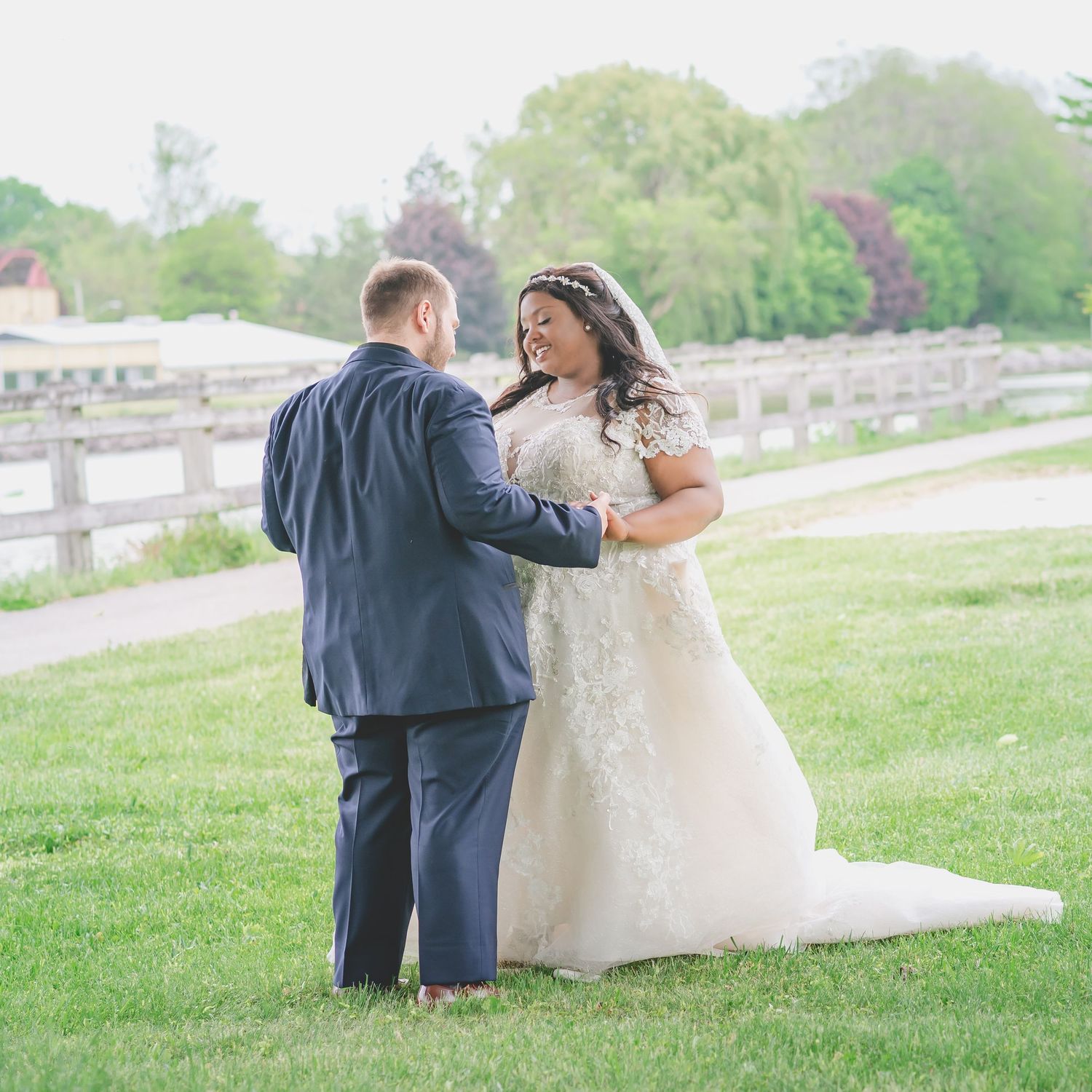 Wedding couple shares a romantic moment on grassy hillside with wooden fence in background.