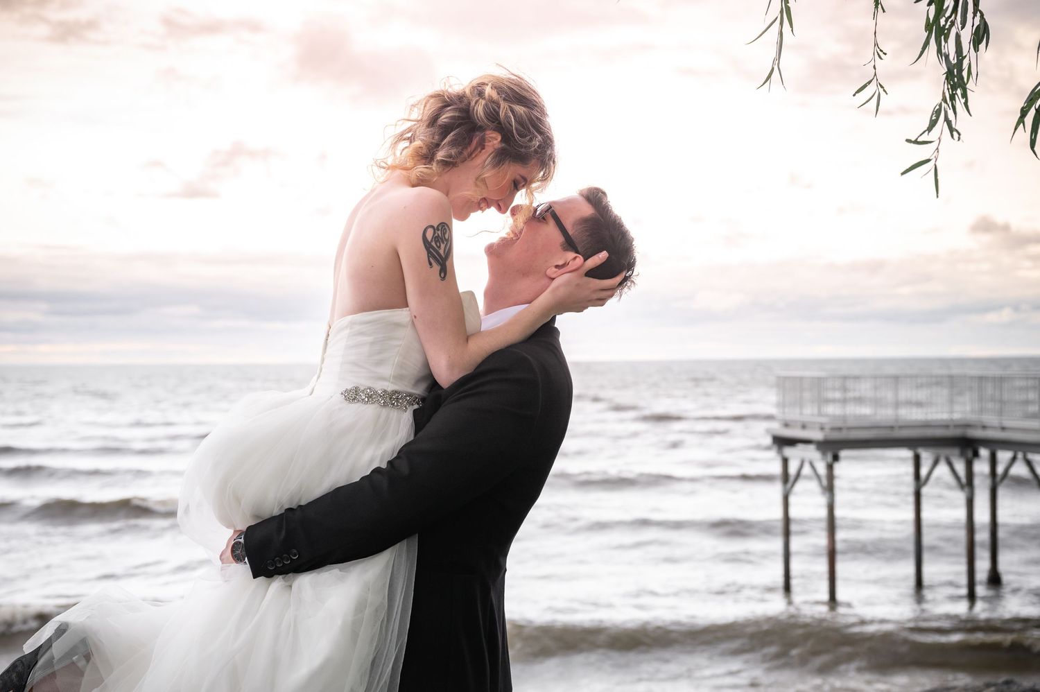 Dreamy sunset beach wedding photo with stylized romantic pose near a pier.