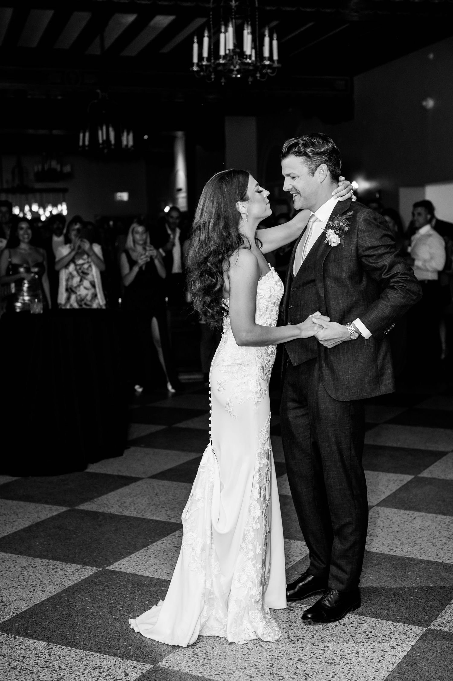 A couple shares their first dance as newlyweds on a checkered dance floor under soft chandelier lighting.