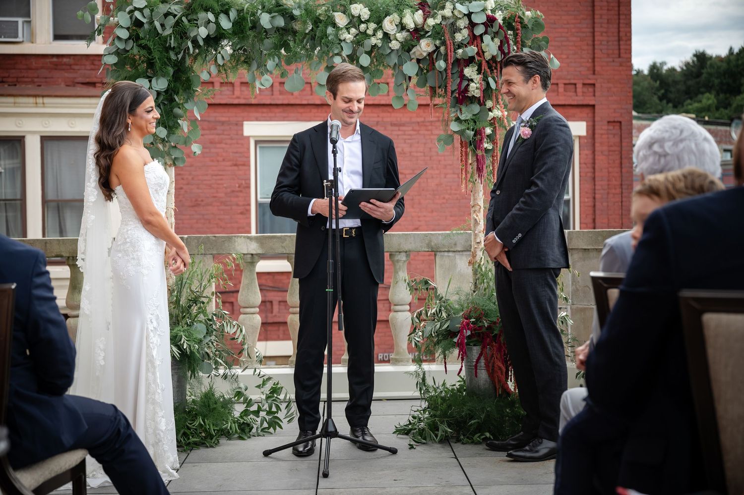 Outdoor wedding ceremony with greenery arch against red brick building.