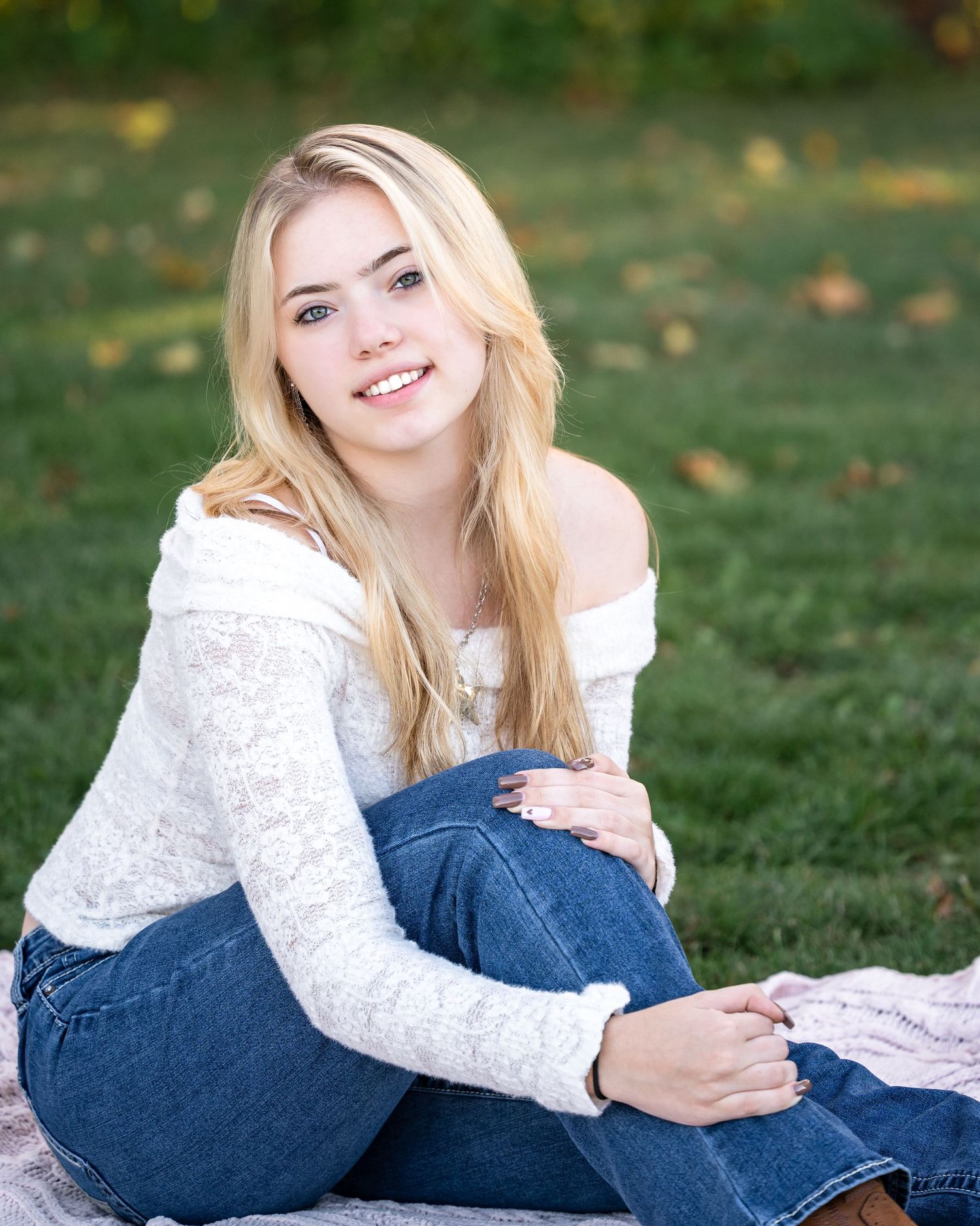 Person in white sweater and blue jeans sitting on grass during autumn season with fallen leaves in background.
