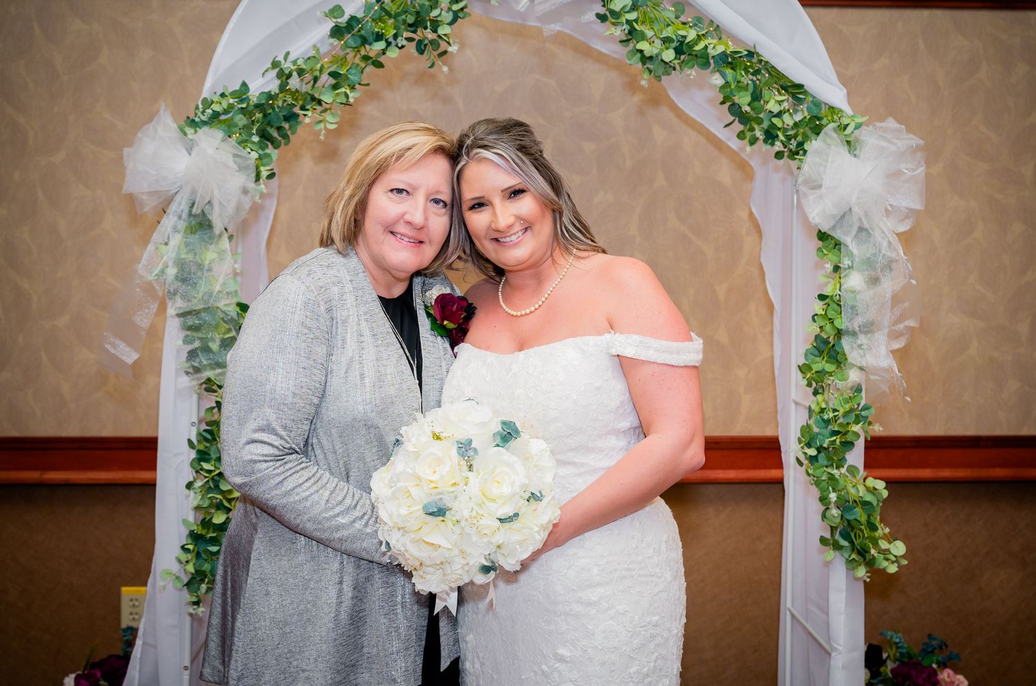 Two people pose under a decorated wedding arch with white flowers and greenery.