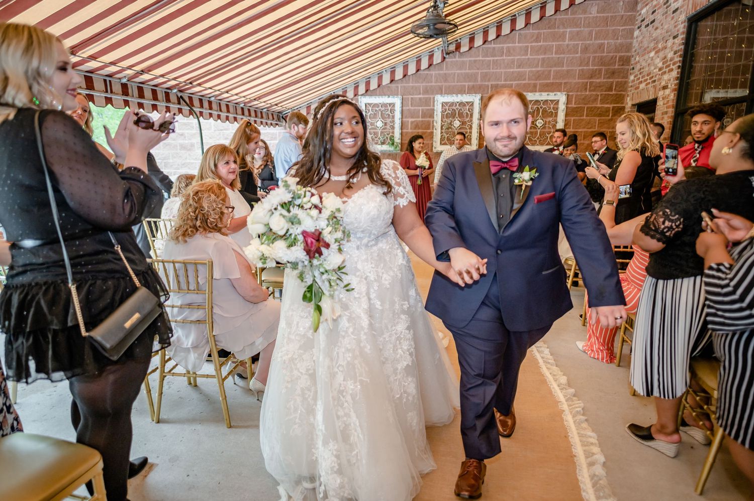 Wedding celebration with guests cheering as the newlyweds exit under a striped awning.