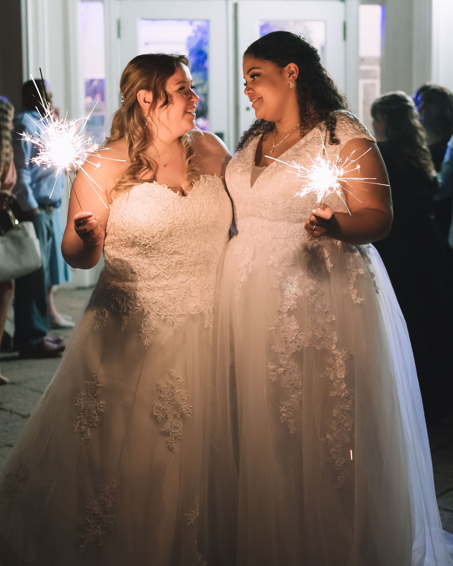 Two brides in white dresses hold sparklers during wedding celebration.