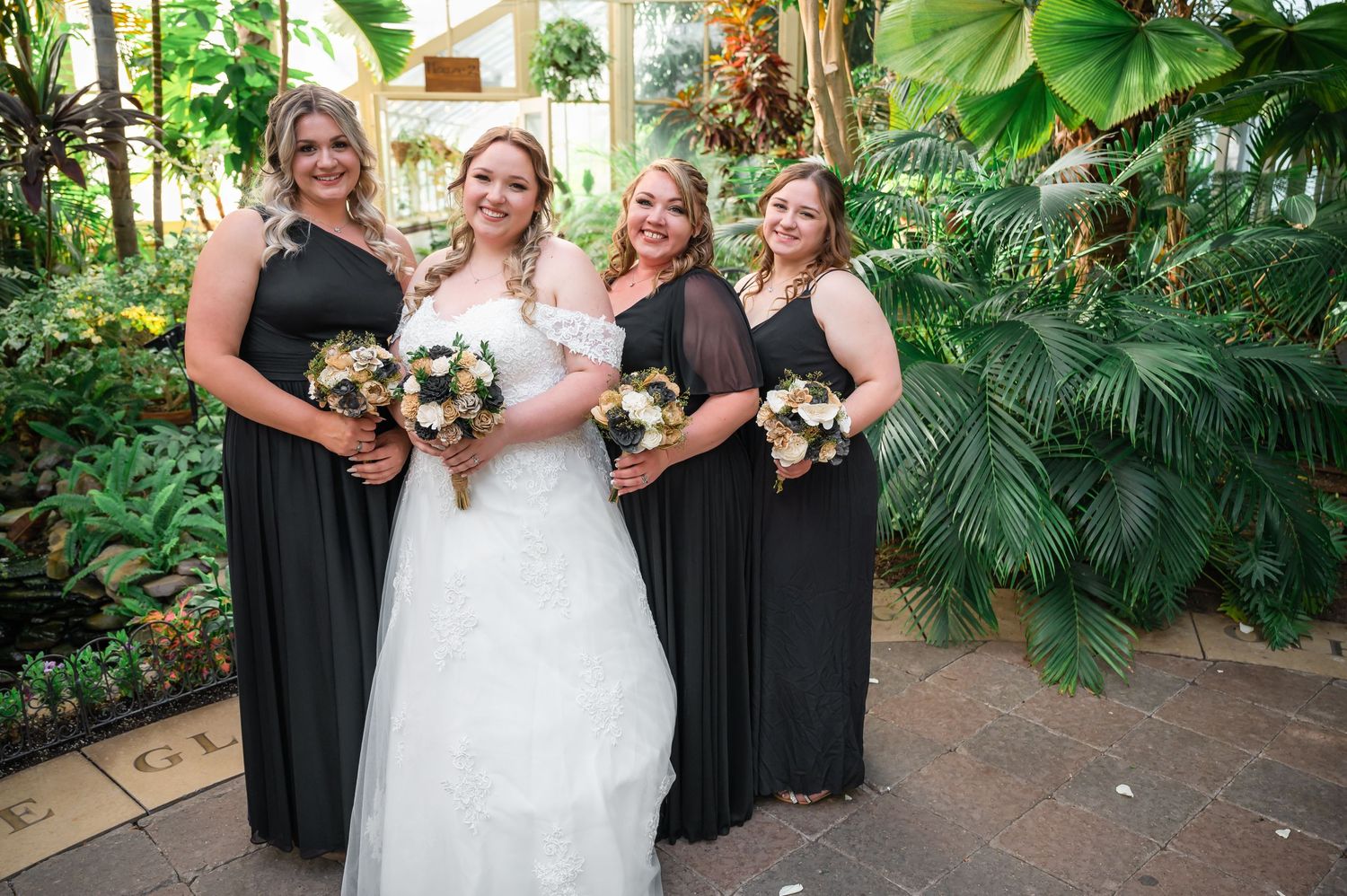 Wedding party poses together holding matching bouquets in a greenhouse setting with tropical plants.