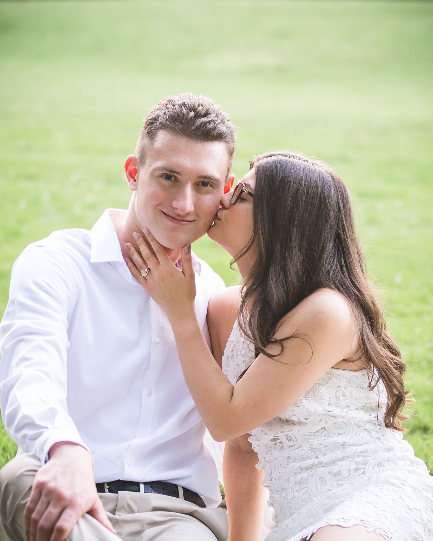 Couple sitting on grass, woman kissing man on cheek during engagement photo shoot outdoors.