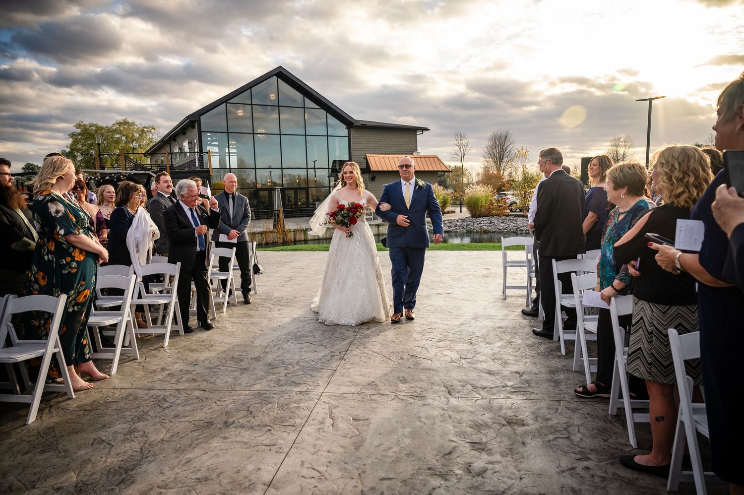 A wedding ceremony takes place outdoors at sunset with dramatic clouds overhead and guests seated in folding chairs.