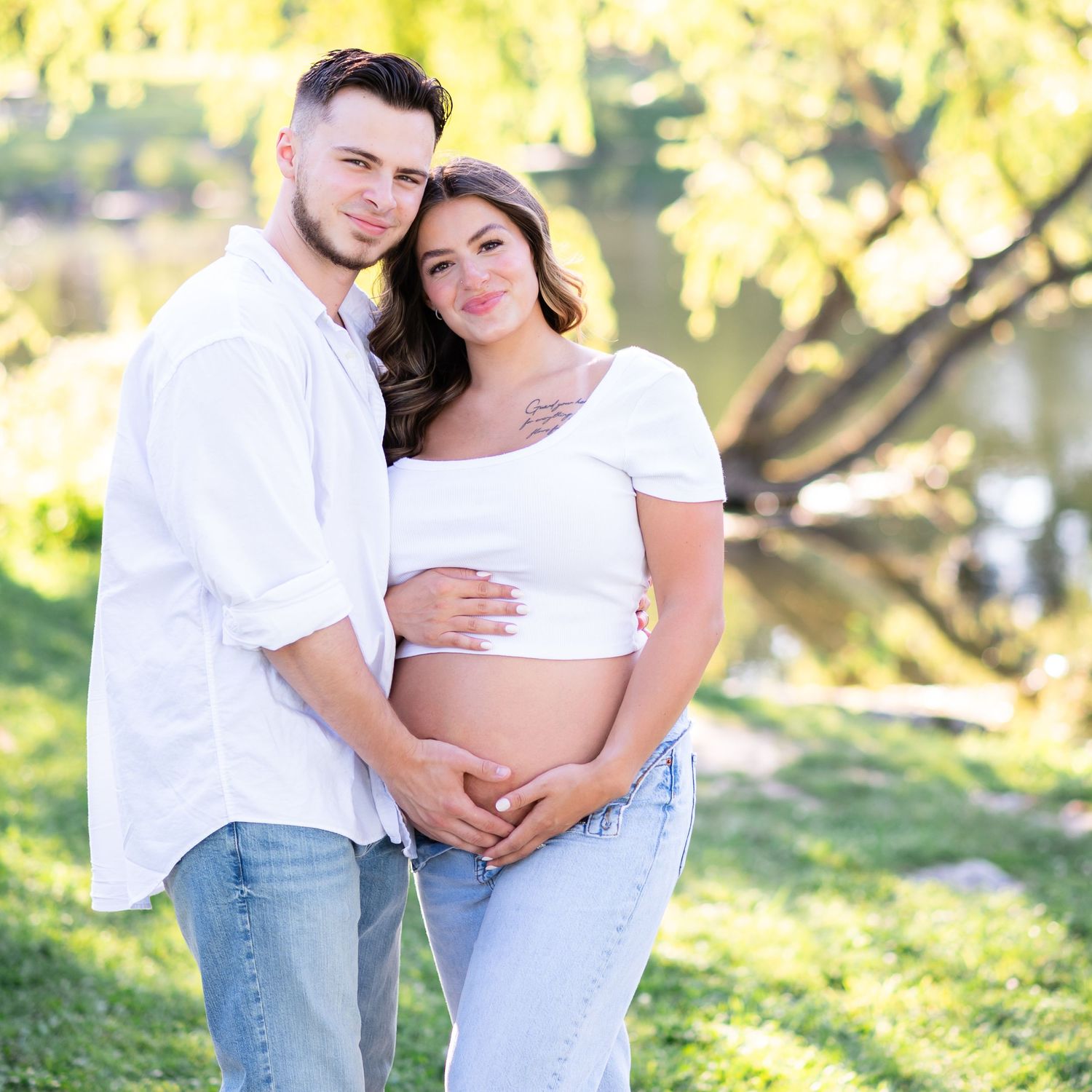 Couple posing for outdoor maternity photo shoot in white outfits, cradling pregnant belly near a serene pond.