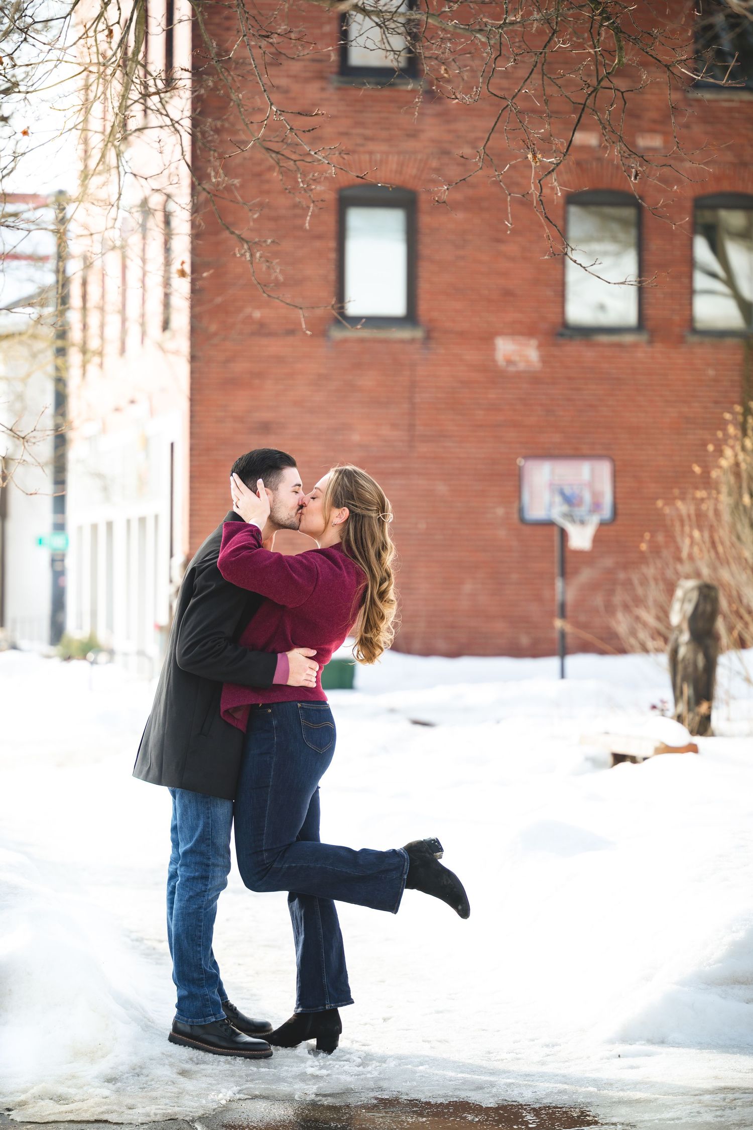 Couple sharing a romantic kiss in a snowy urban setting with a red brick building backdrop during winter engagement photos.