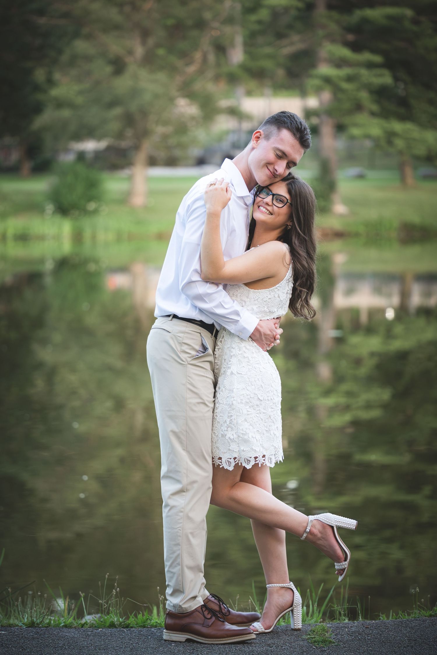 Couple embracing and smiling by a serene pond, woman in white lace dress, man in white shirt and khaki pants.