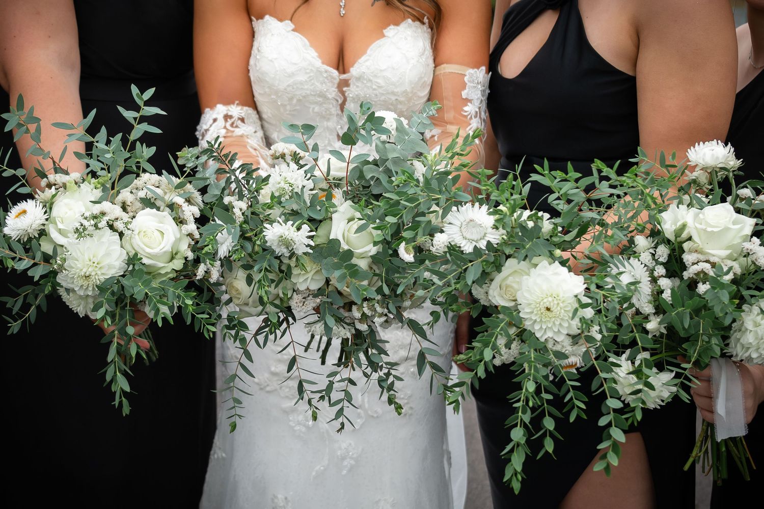 A row of wedding party members holding elegant white and green floral bouquets against black attire.