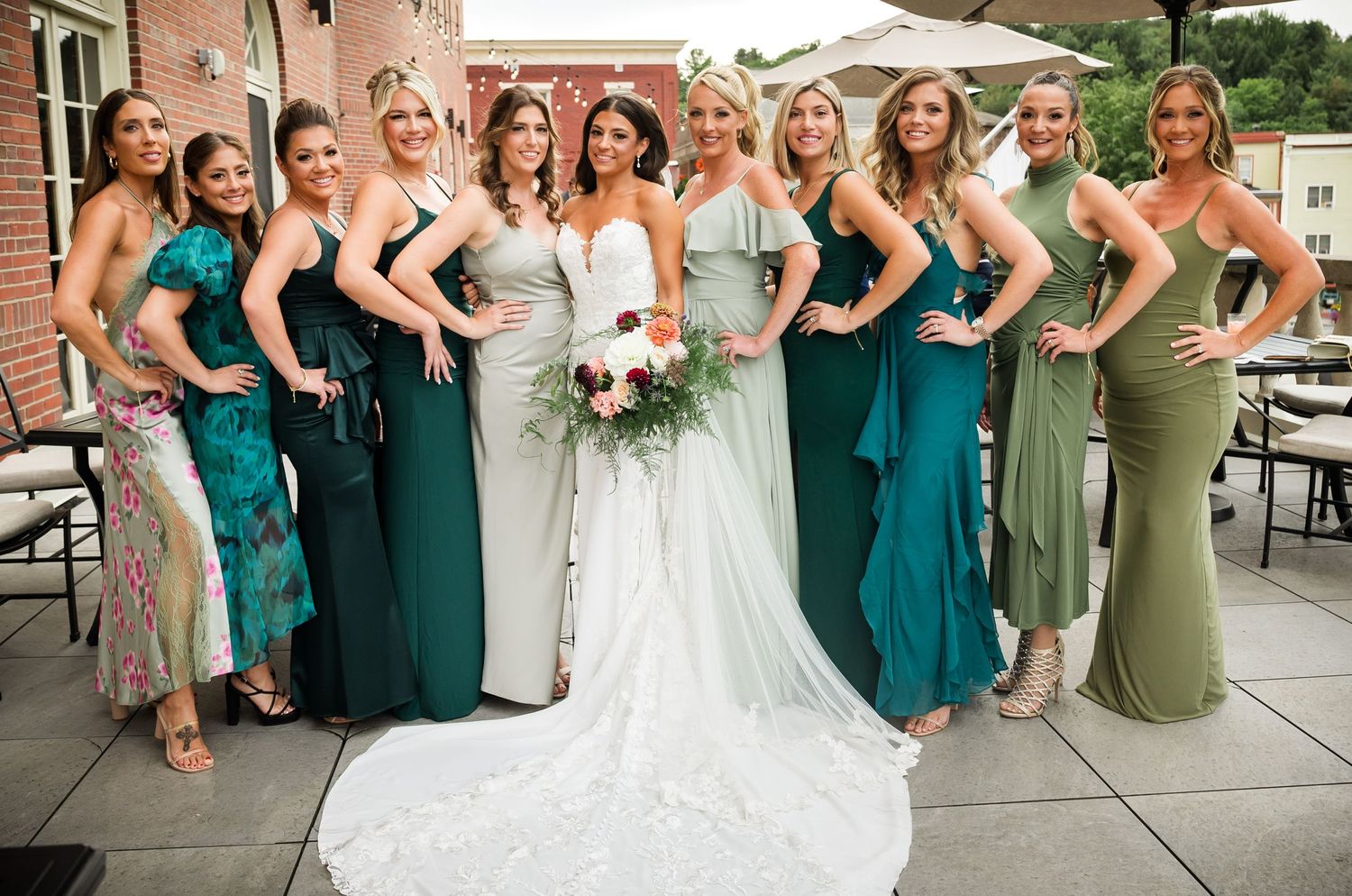 Bridesmaids in elegant teal and sage green dresses pose together on a rooftop terrace.