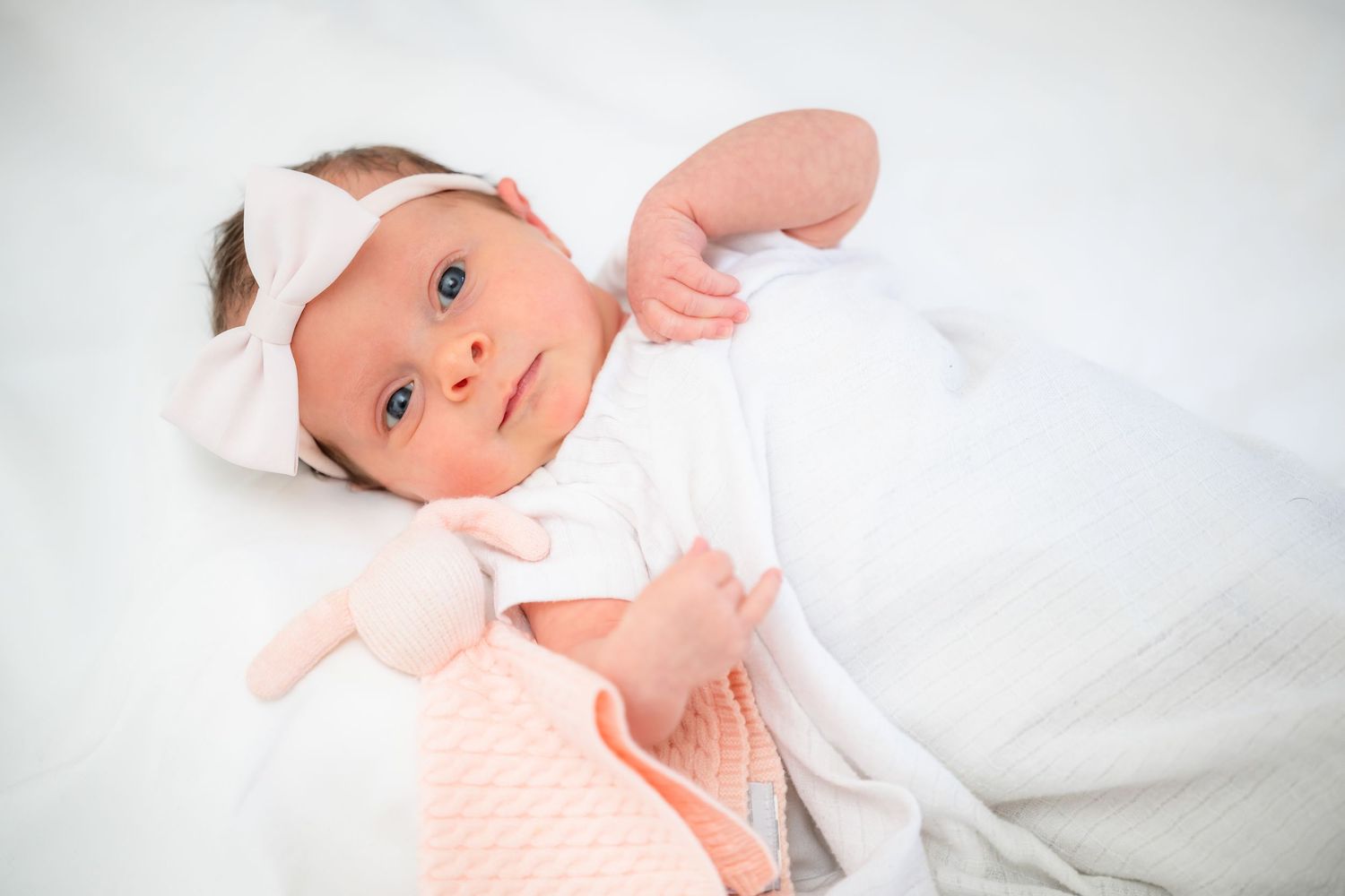 Newborn baby wearing white clothing lying on soft white bedding.