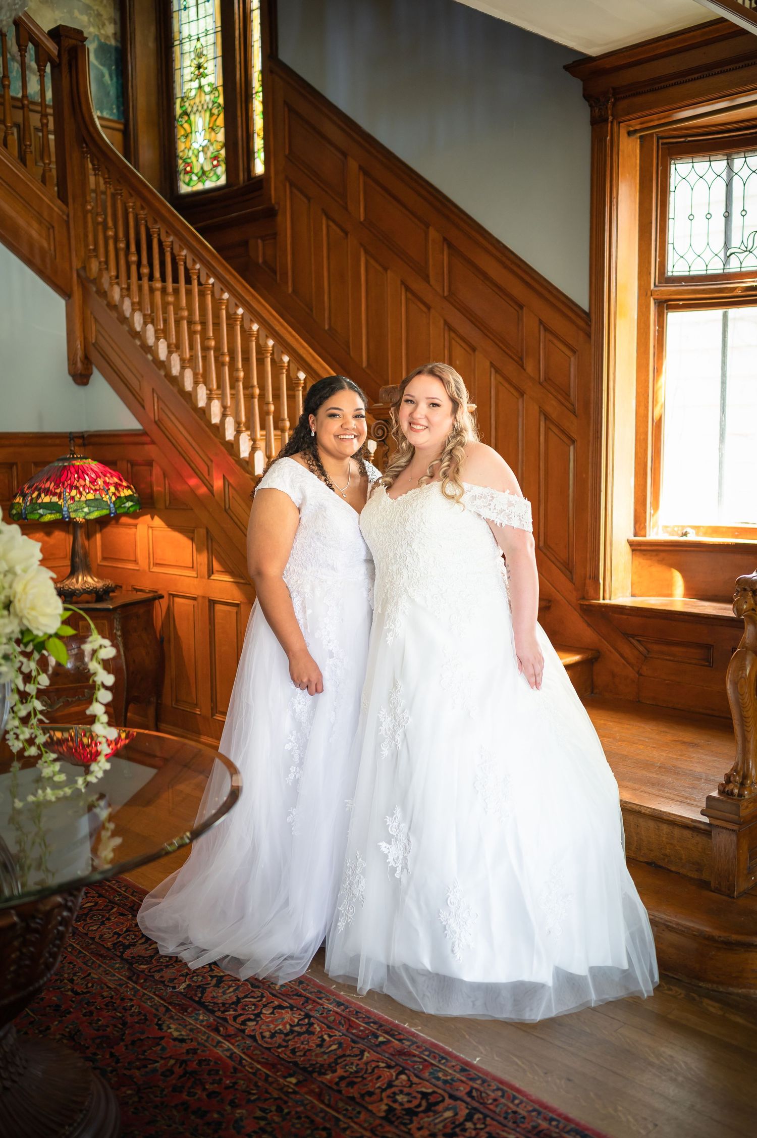 Two people in white wedding gowns stand on wooden staircase surrounded by elegant decor.