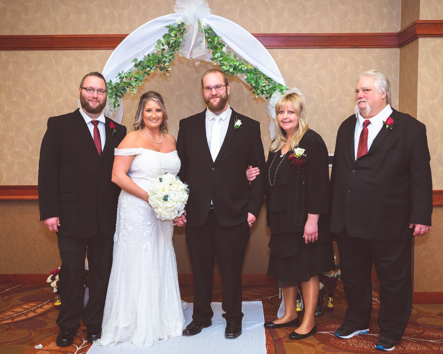 Formal wedding party group photo under white arch with greenery in an indoor venue setting.