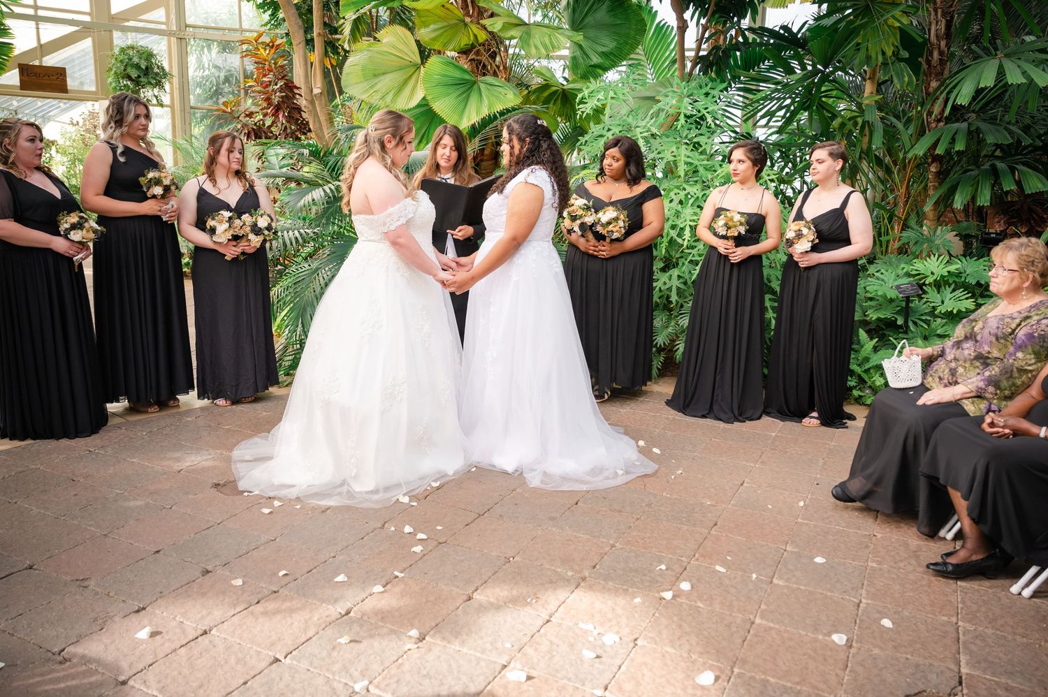 Wedding ceremony with two brides in white dresses surrounded by bridesmaids in black gowns.