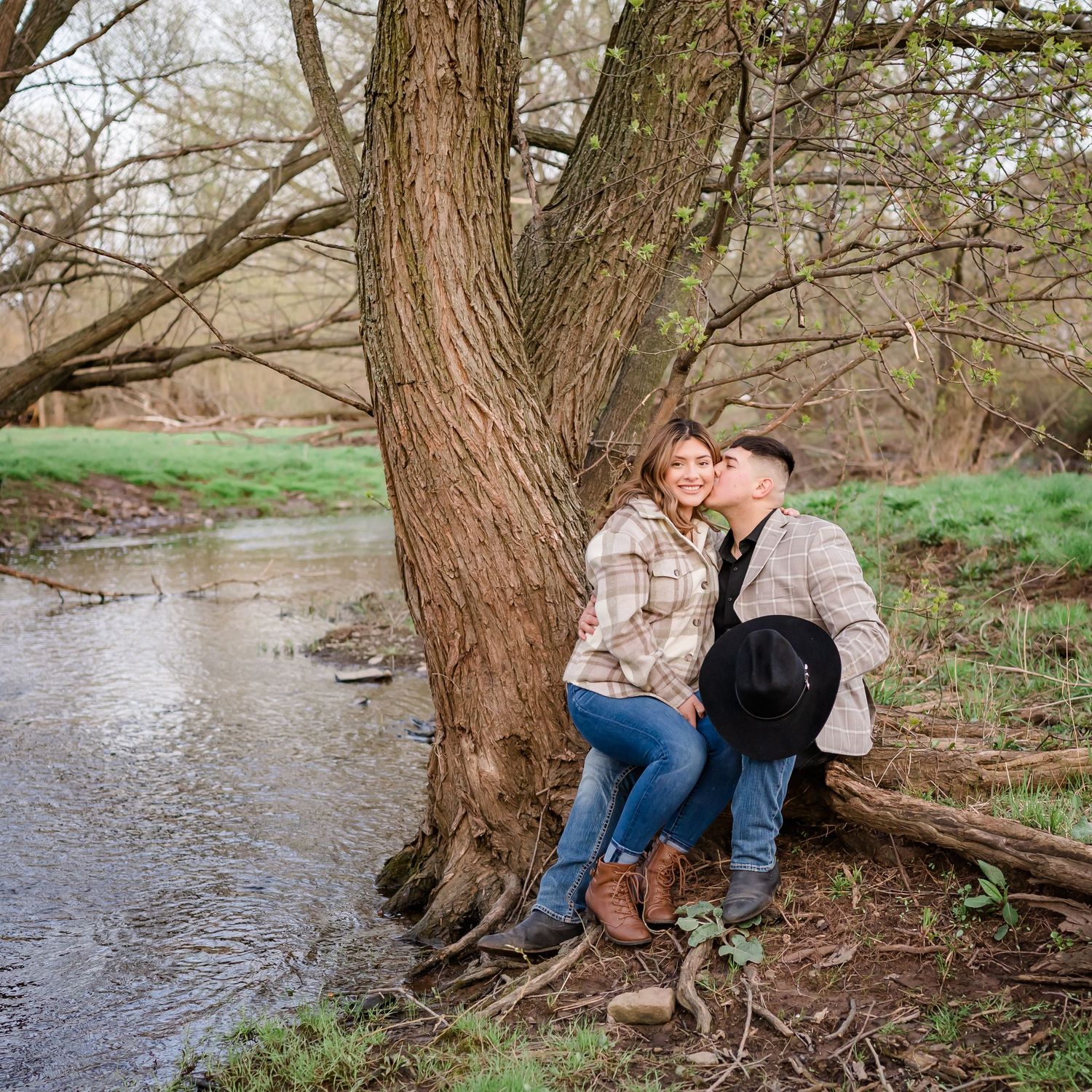 A couple sharing an intimate moment while sitting on a large tree trunk by a stream.