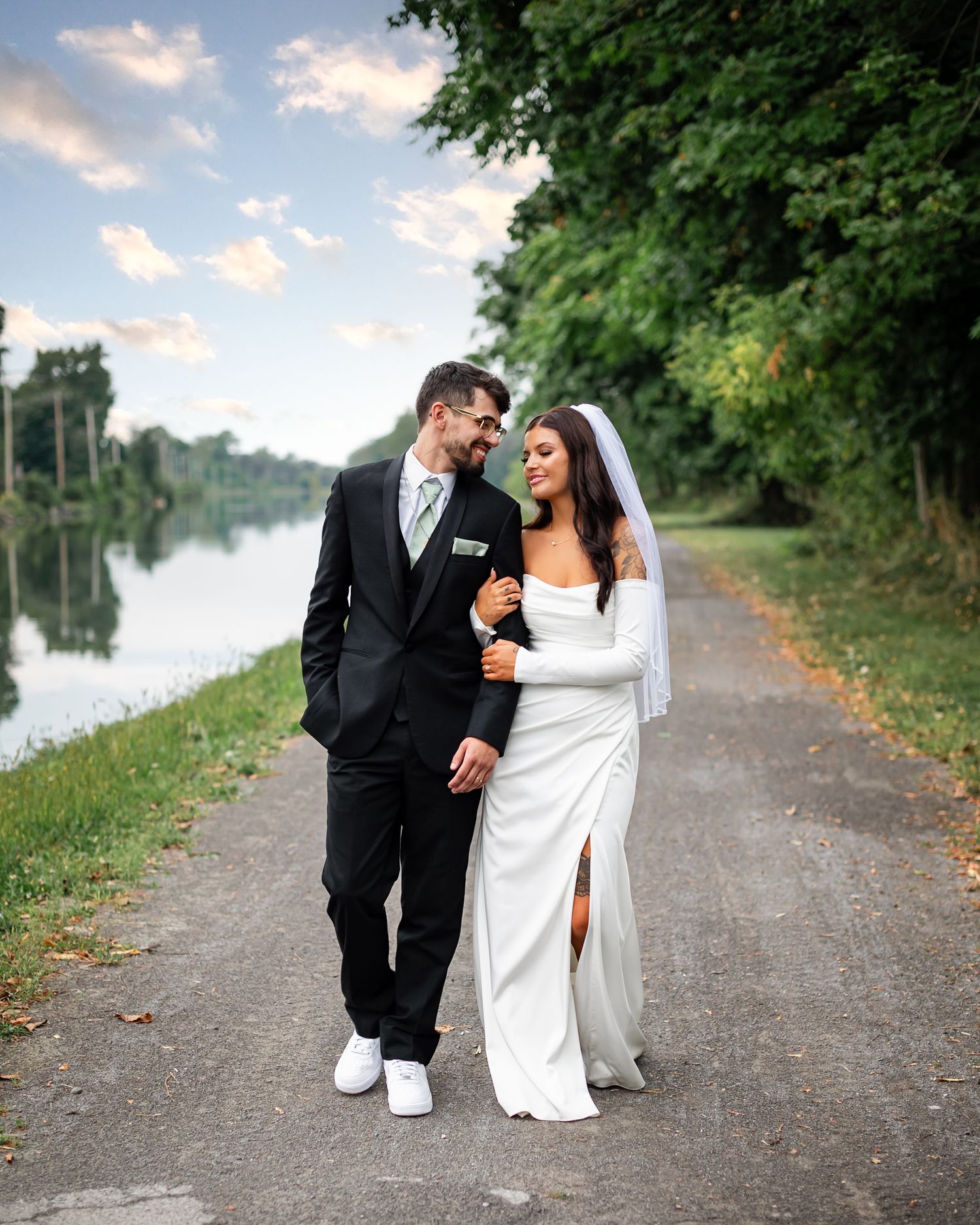 A couple in wedding attire shares a joyful moment walking together on a scenic path by the water.