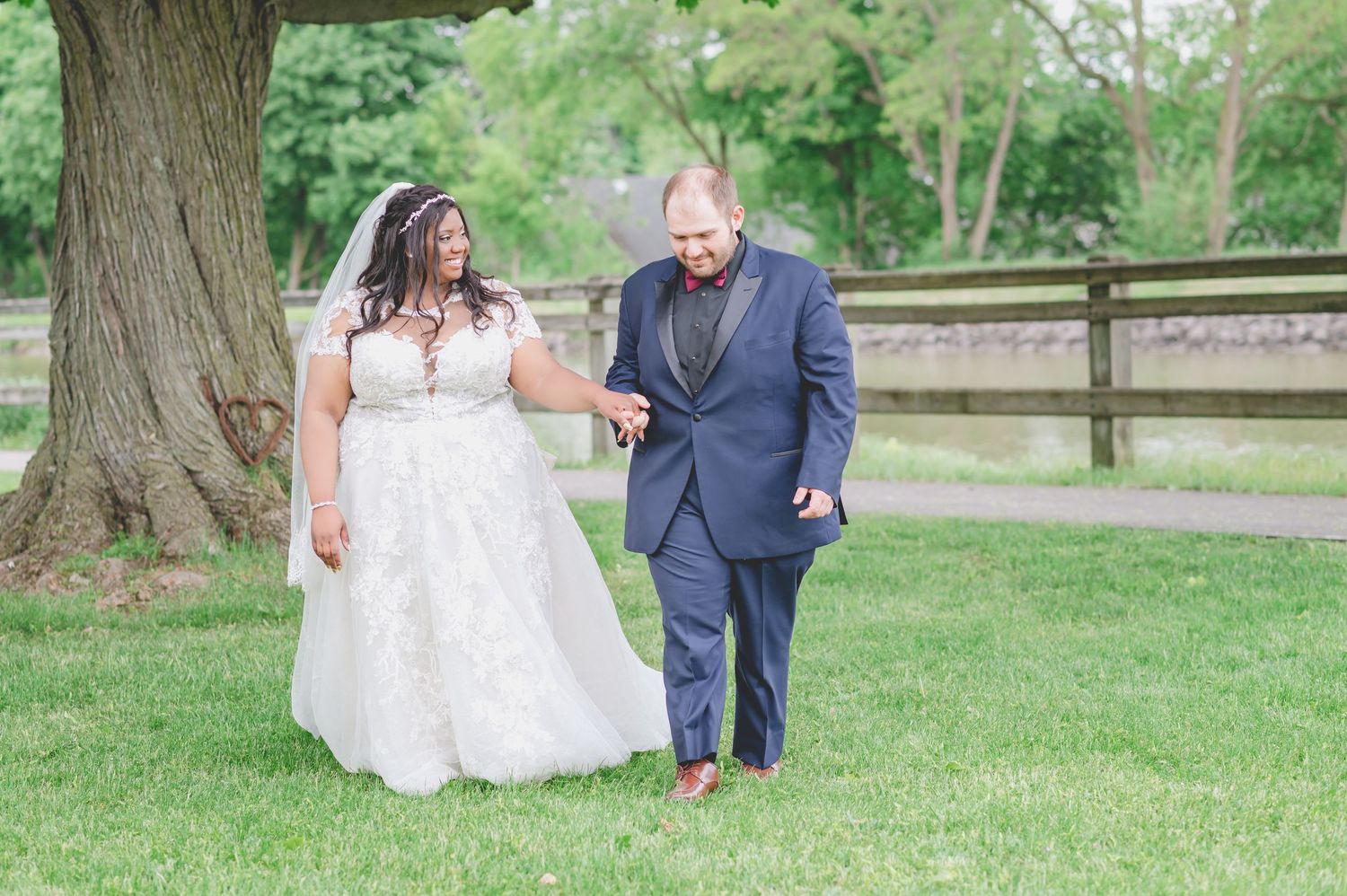 Newlyweds walk together on green lawn by large tree, sharing a lighthearted moment.