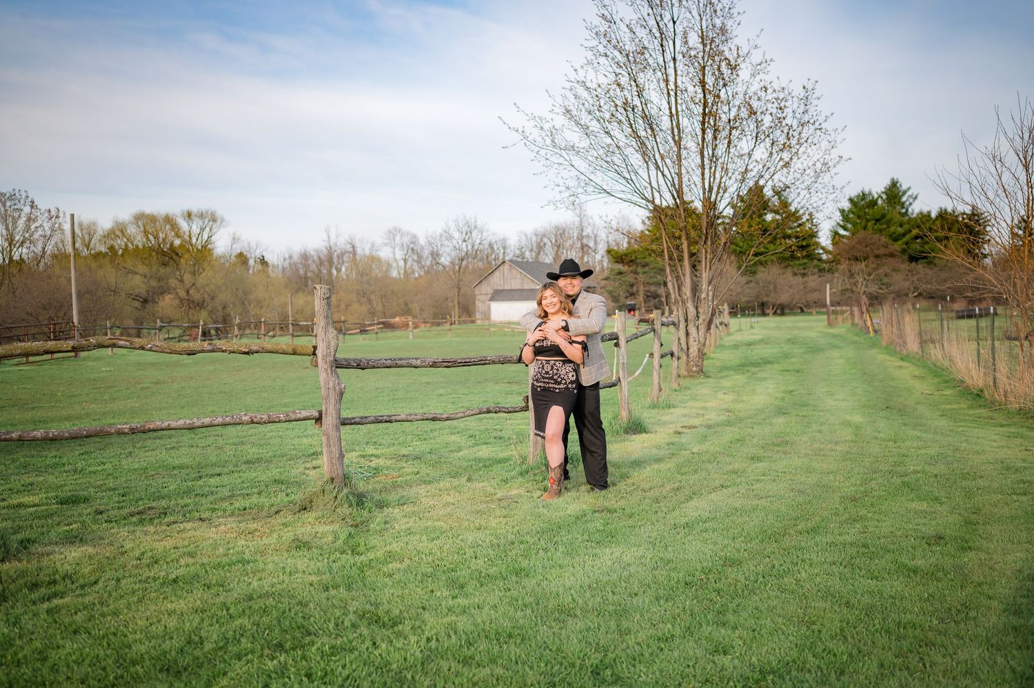 Couple embracing in green countryside with fence posts and trees in background.