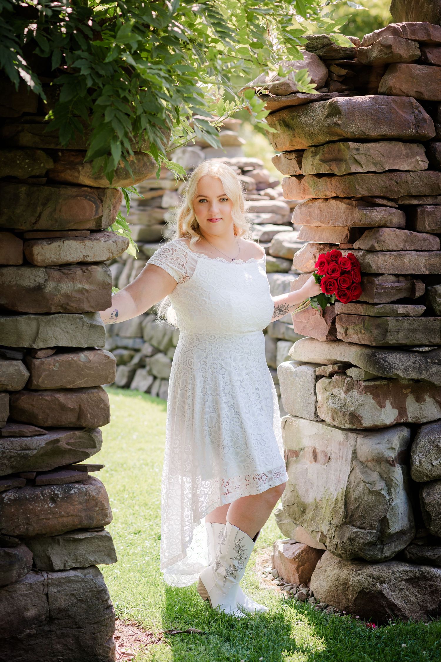 Figure in white lace dress poses in stone garden archway holding red roses.