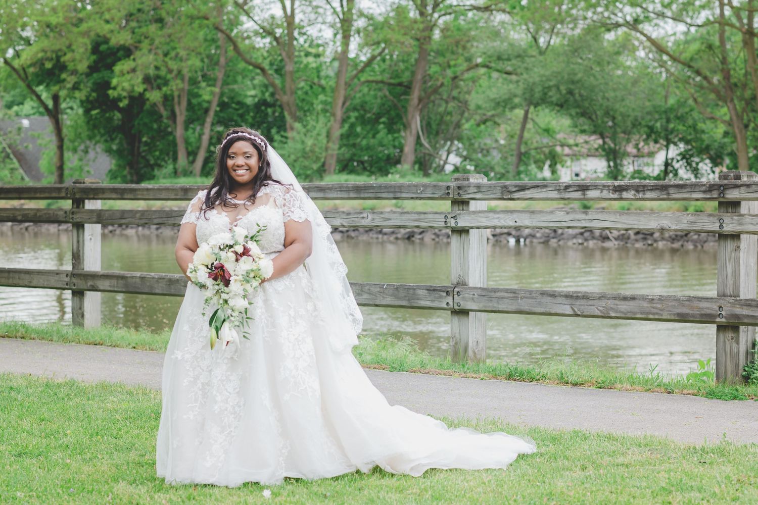A bride in a white lace gown and veil poses outdoors near a wooden fence with her bouquet.