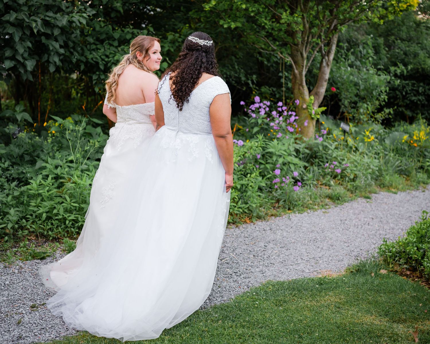 Couple in white wedding dresses walks together along garden path.