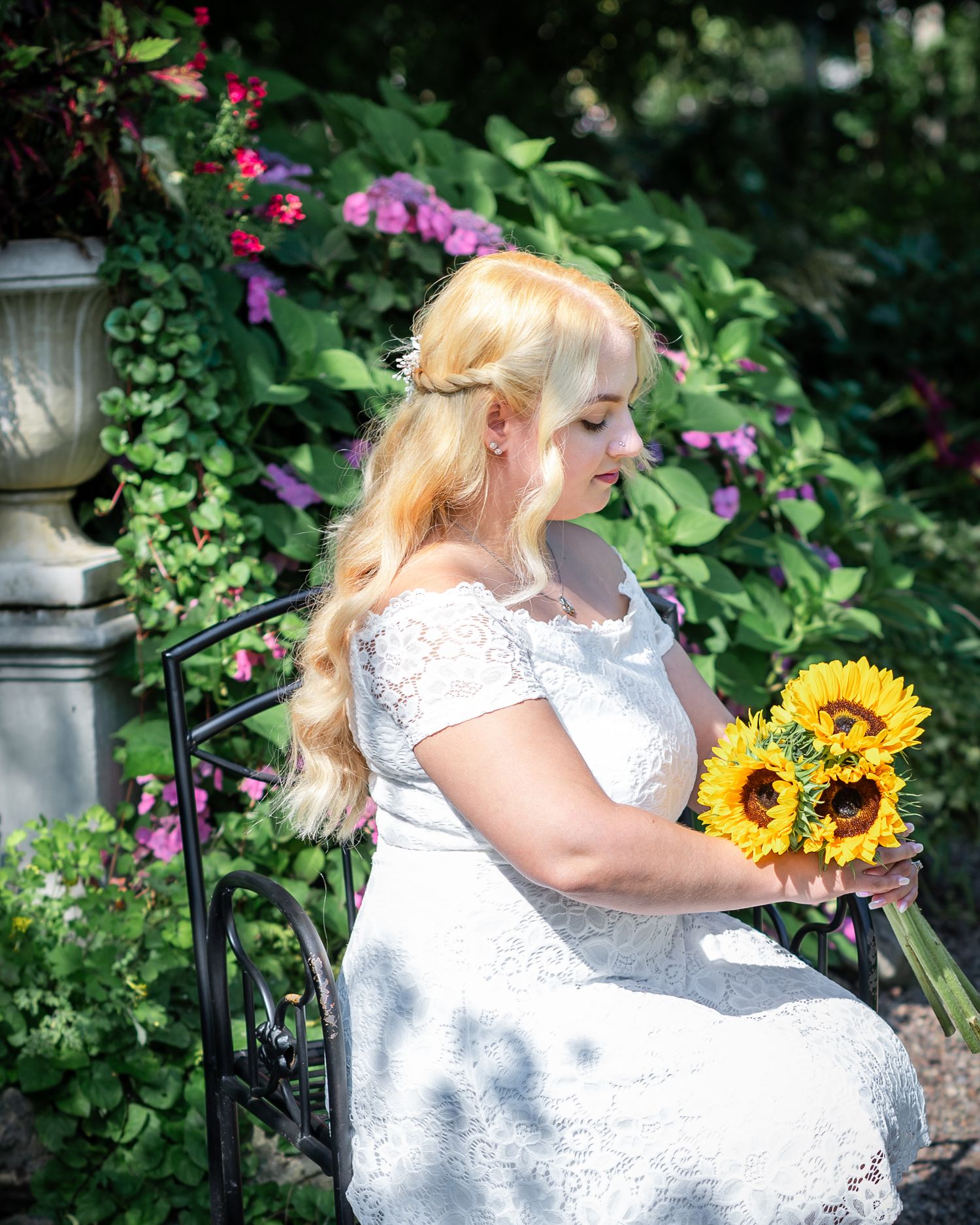 A bride in white dress poses with sunflowers in a lush garden setting with purple flowers.