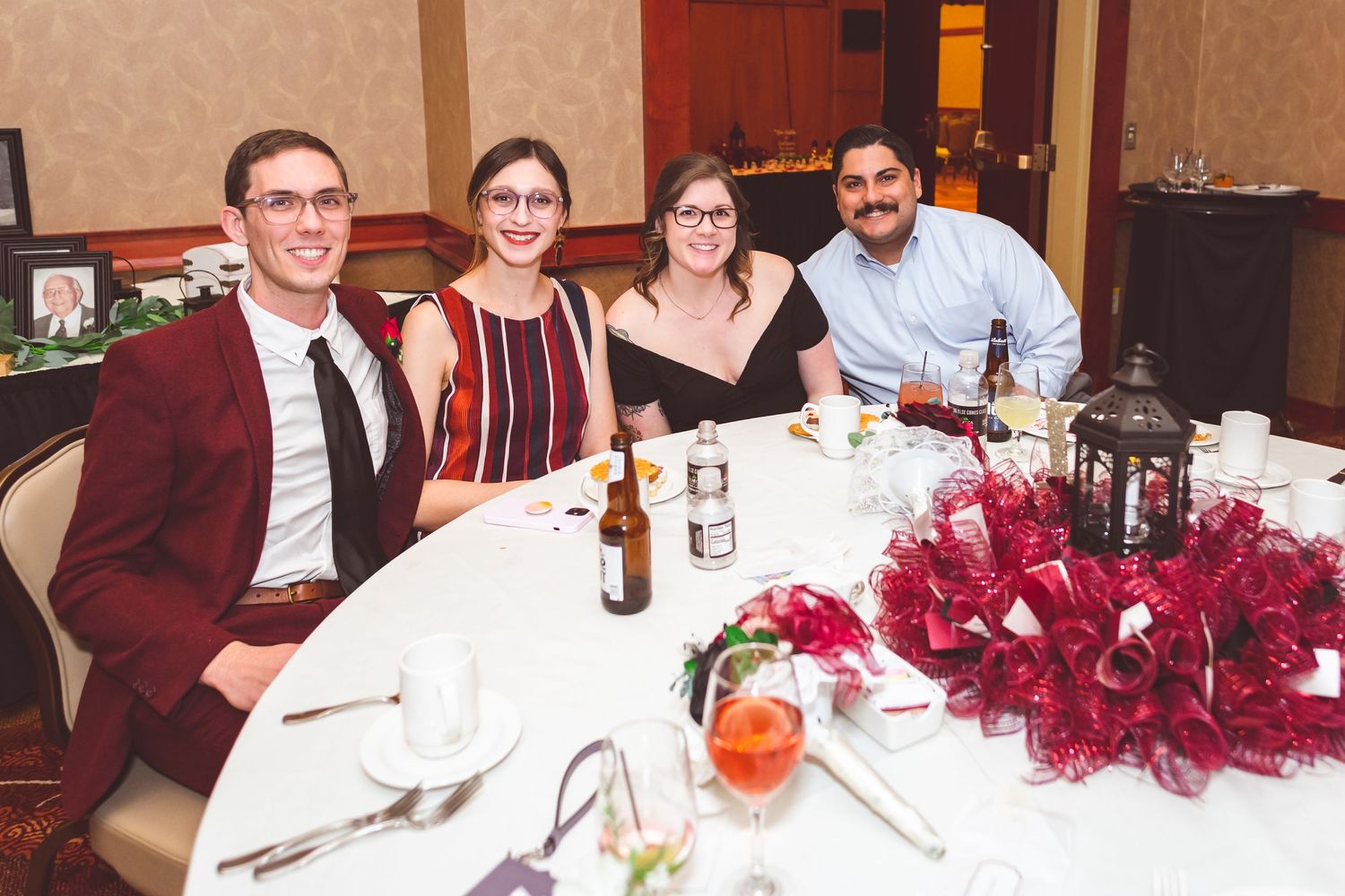 Wedding reception guests gather around table with red floral centerpiece.