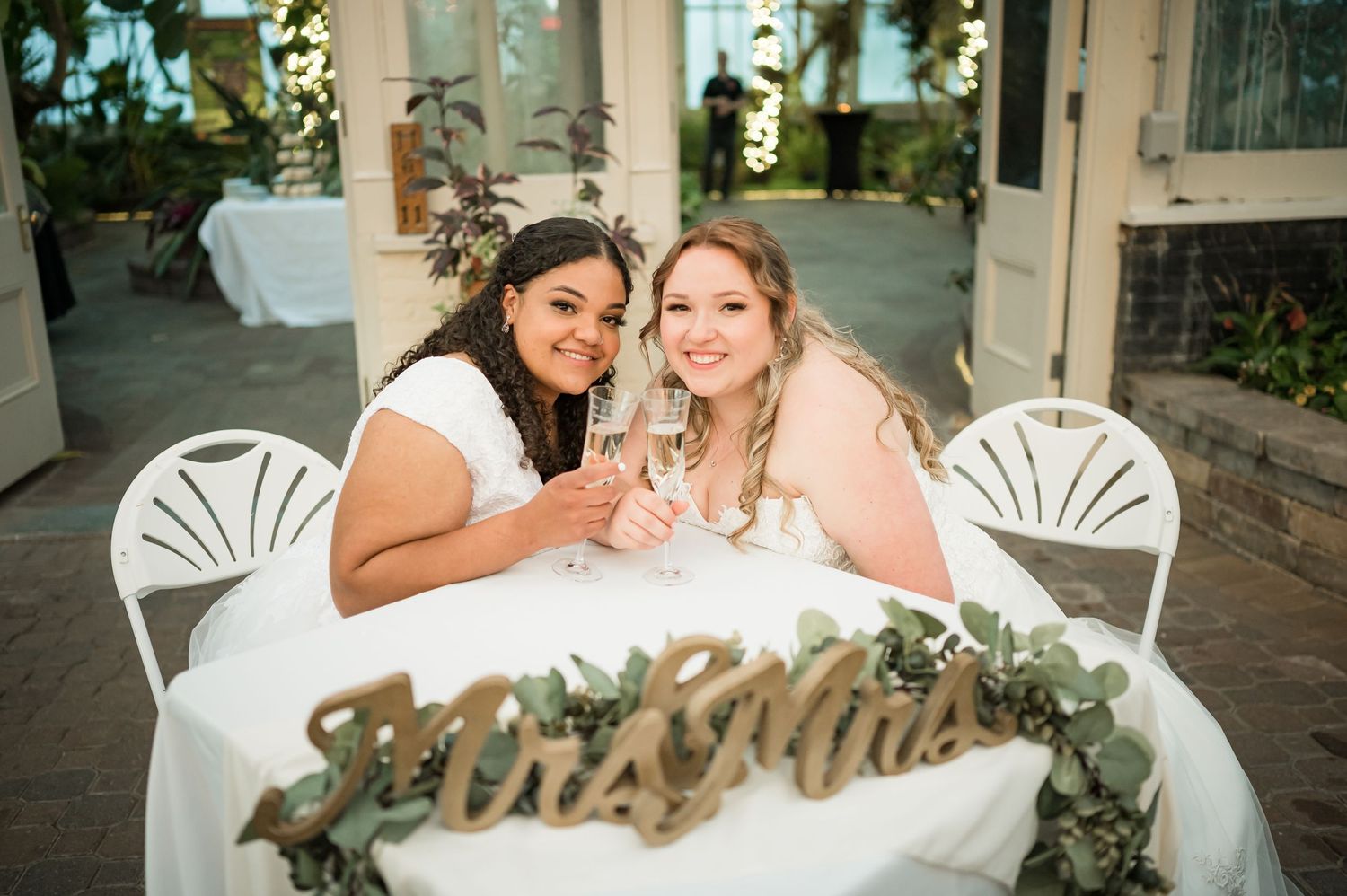 Two brides share champagne while sitting at decorated table with greenery.