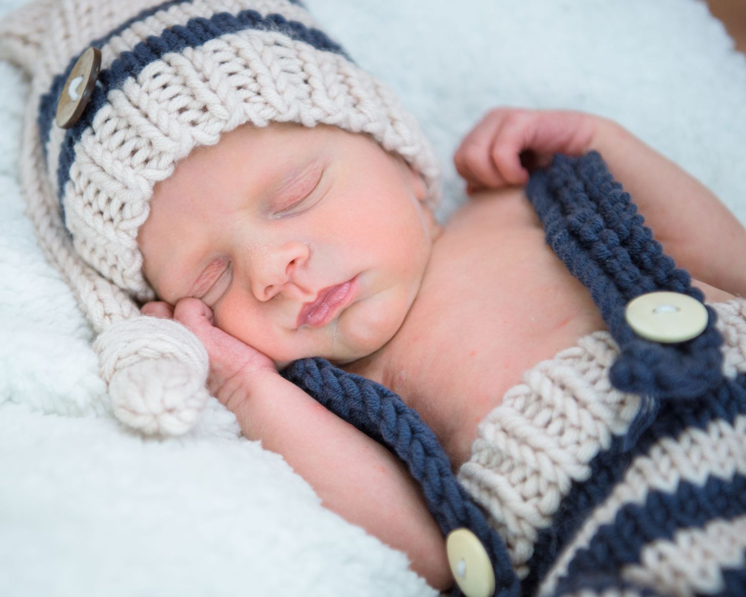 A sleeping baby wearing a white knit hat and navy sweater lies peacefully on soft bedding.