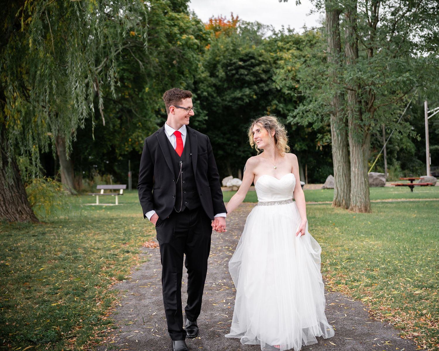 A couple walks along a tree-lined path, the bride in a flowing white gown and groom in classic black suit.