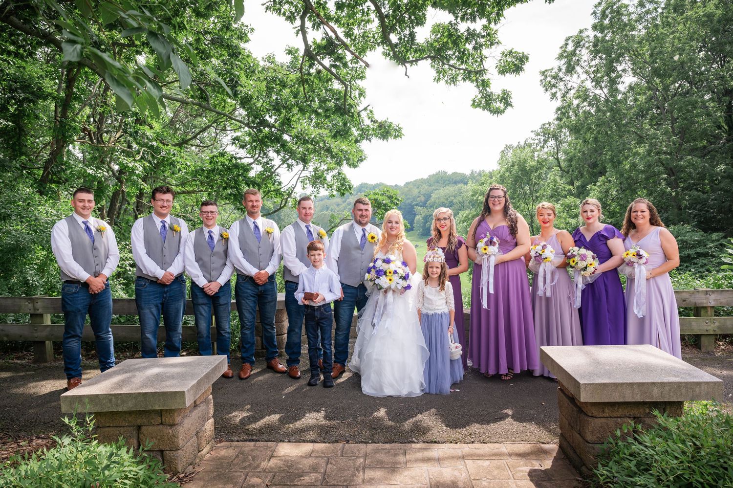 Full wedding party in purple dresses and gray vests lined up on stone terrace for formal group photo.