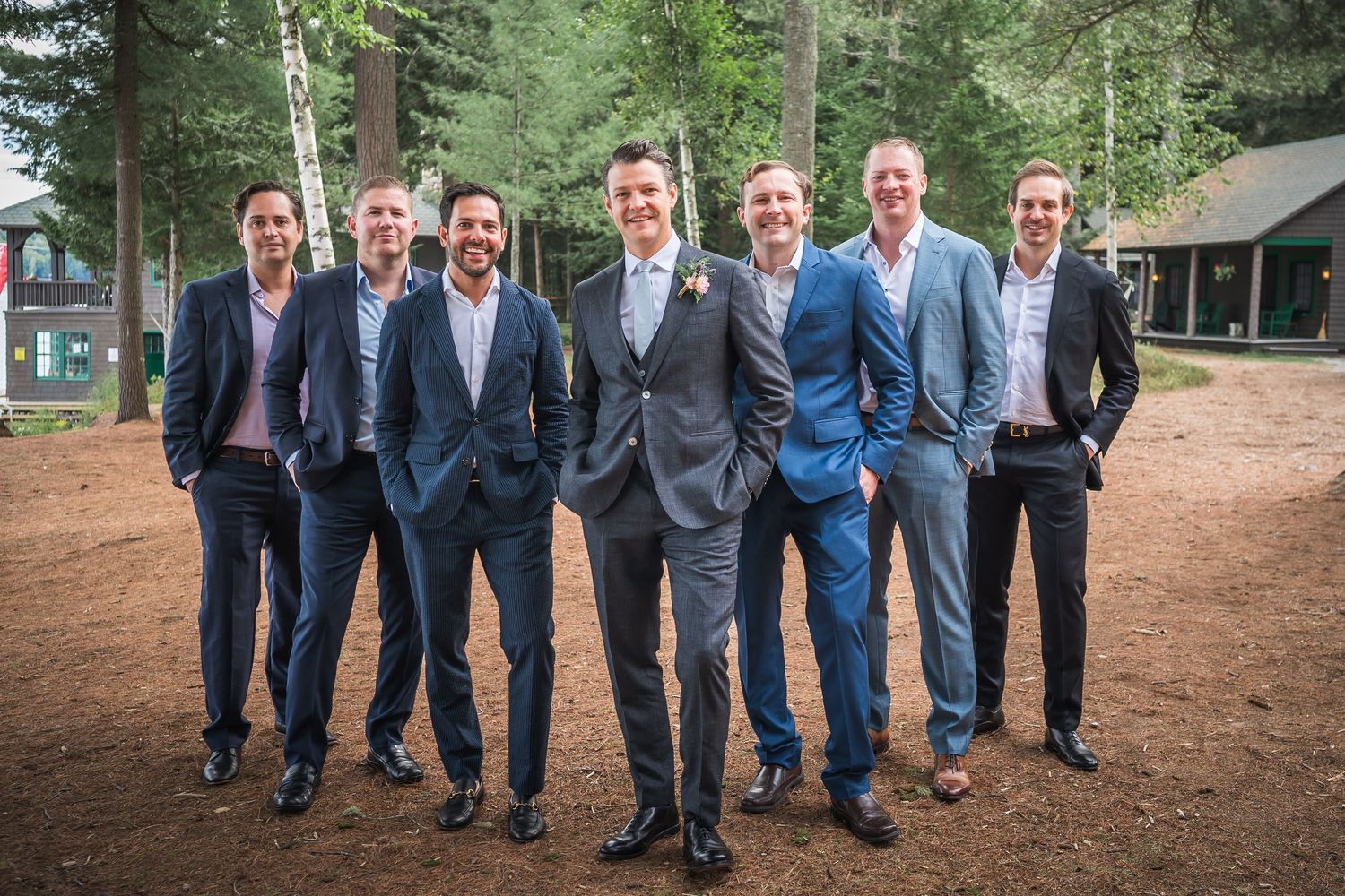 Groomsmen in dark suits pose together on rustic outdoor grounds.