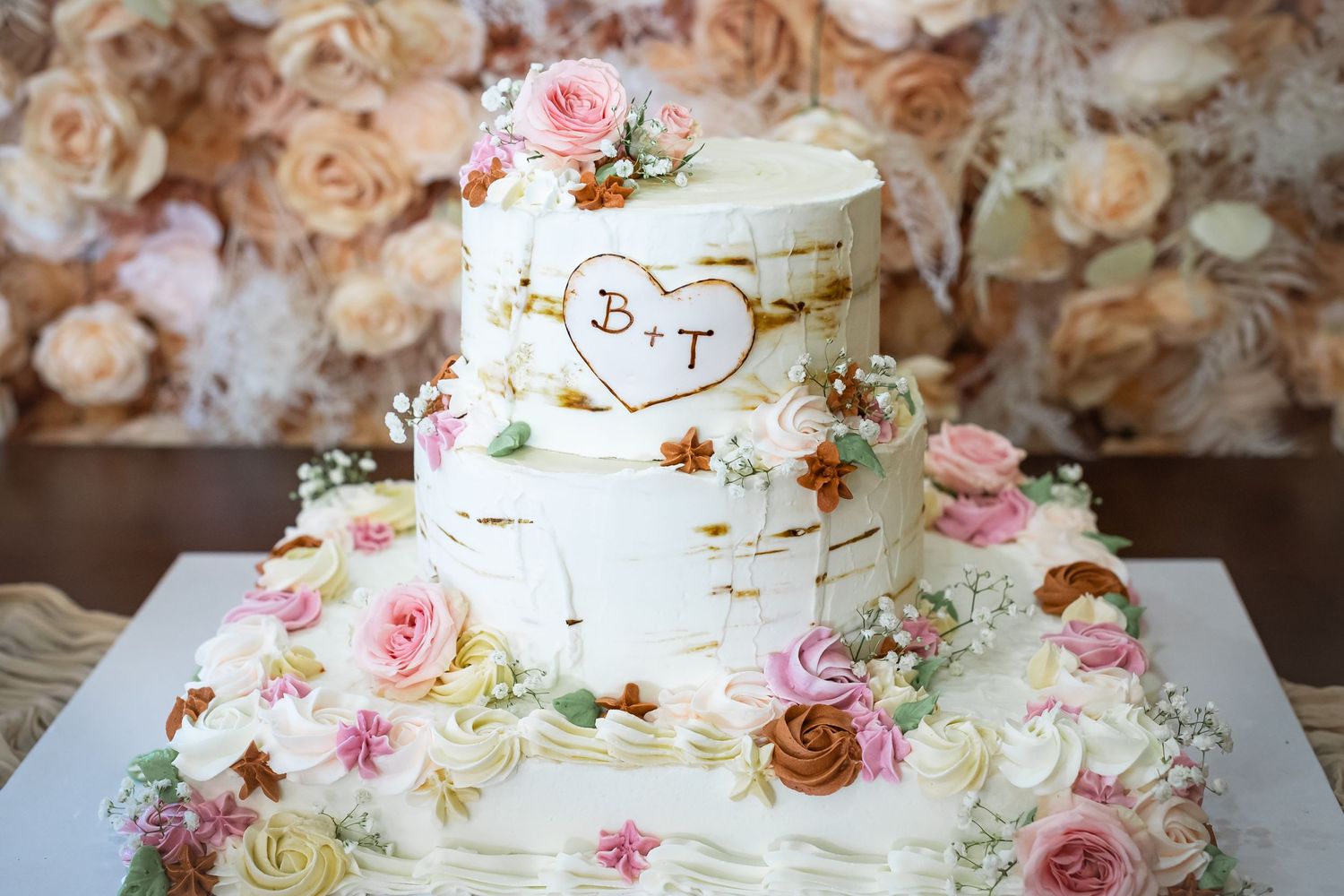 Three-tiered white wedding cake decorated with pink roses and gold accents on a floral background.