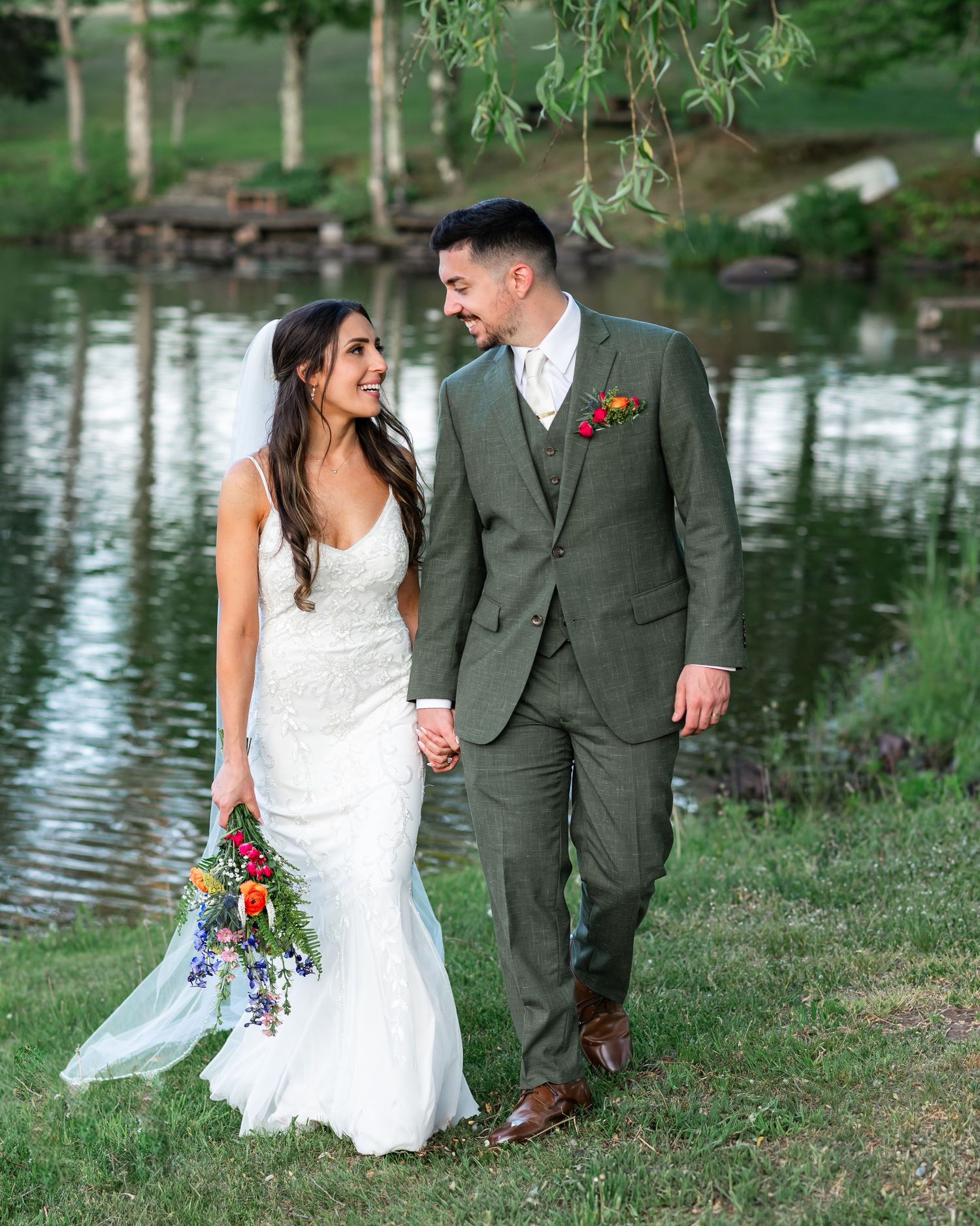 A wedding couple walks together on a grassy path by a serene pond, with the bride in white and groom in gray.