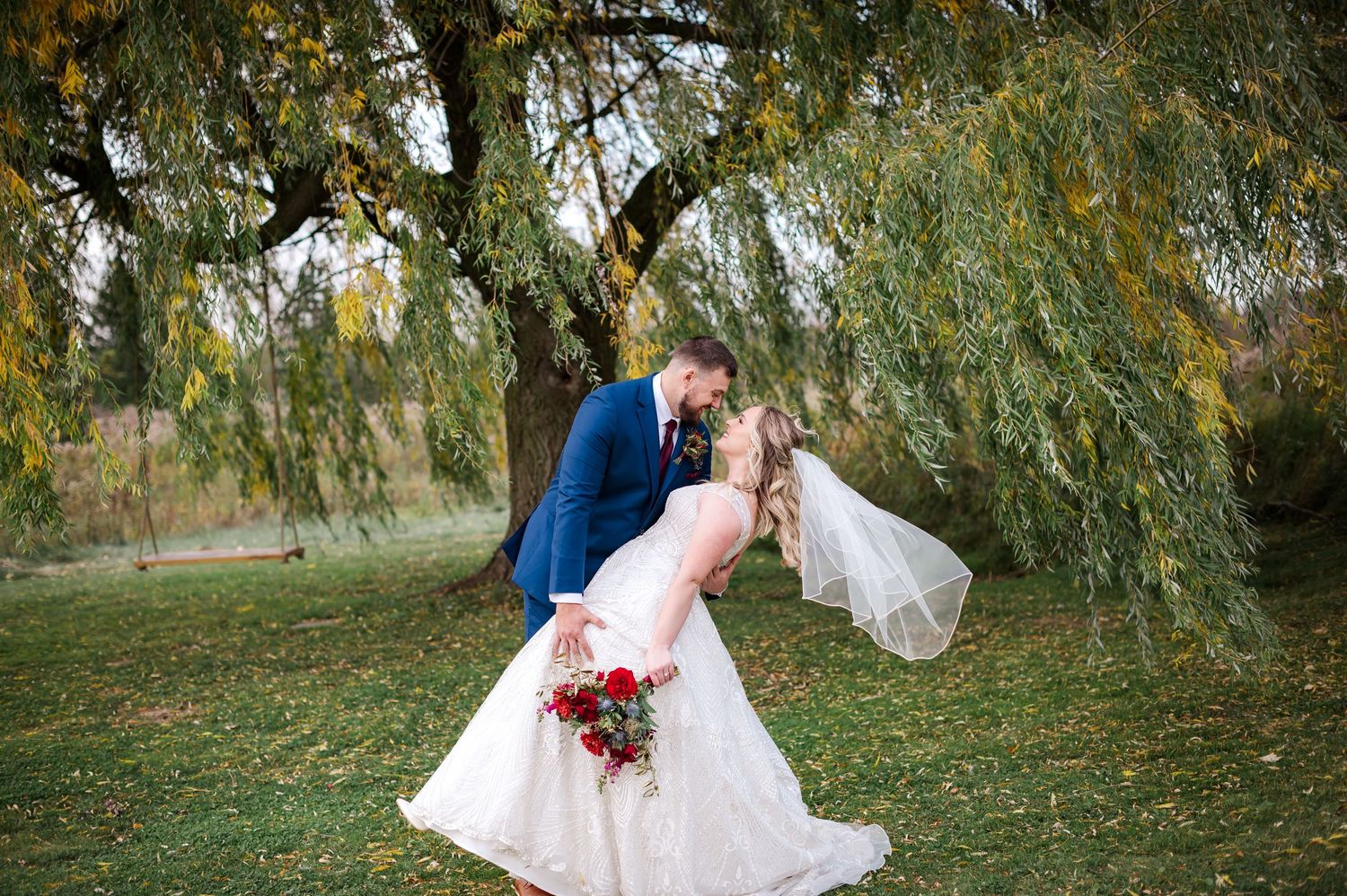 Bride and groom share romantic dip kiss under weeping willow tree, bride holding red bouquet in autumn outdoor wedding.